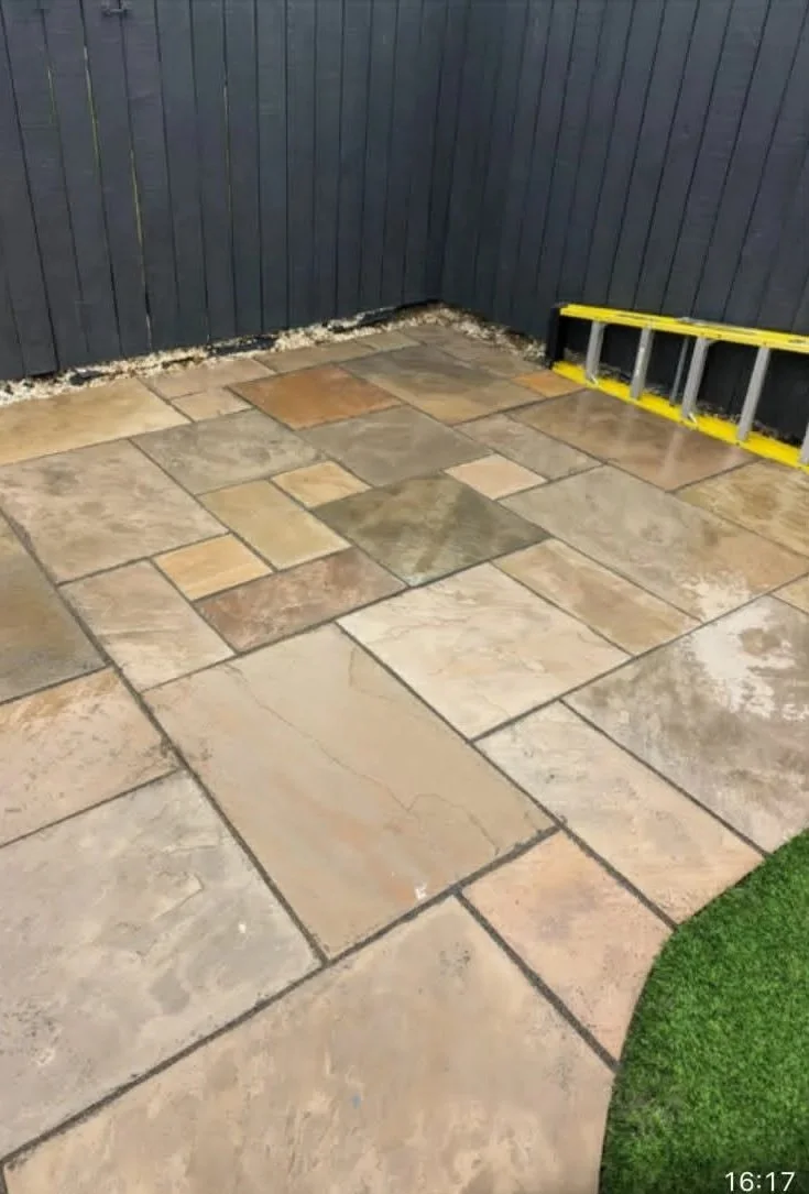 Backyard patio with large stone tiles, a black wooden fence, a yellow ladder, and artificial green grass in the corner.