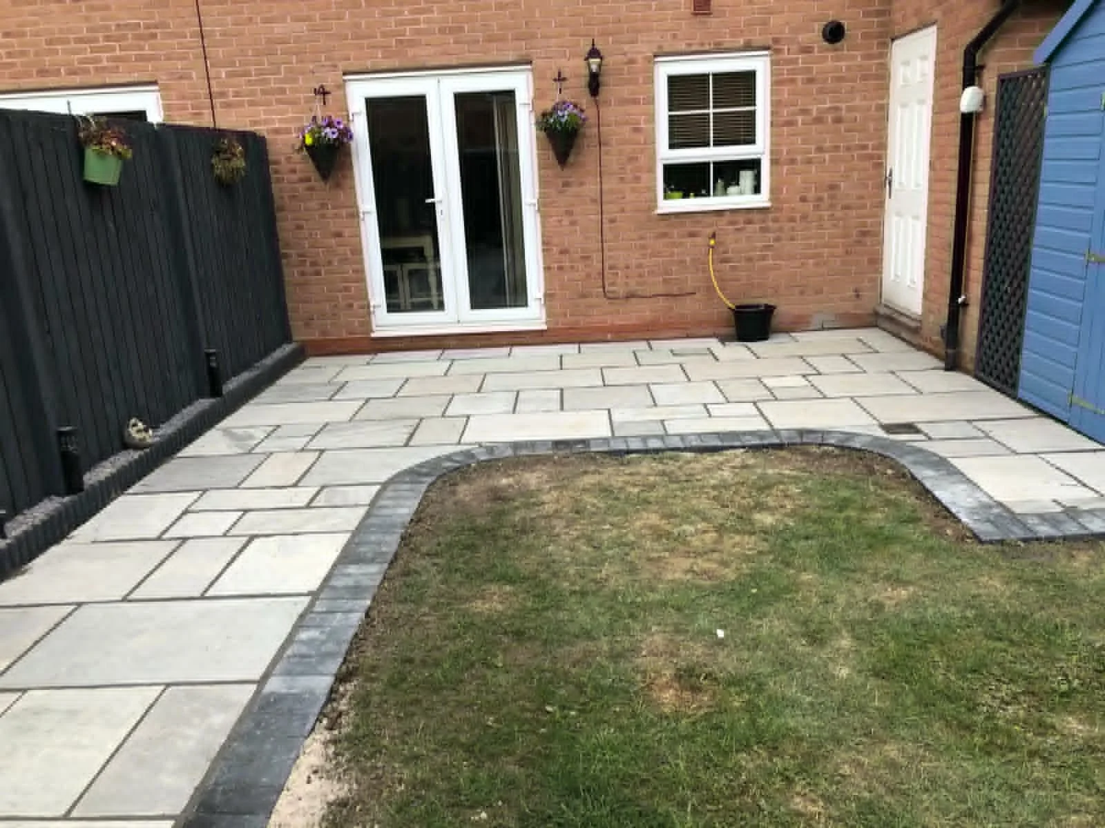 A backyard patio with stone pavers along the house and a grassy area in the center. The house has brick walls, two windows, a sliding glass door, and a white door. There are hanging flower baskets and an outdoor light fixture on the wall.