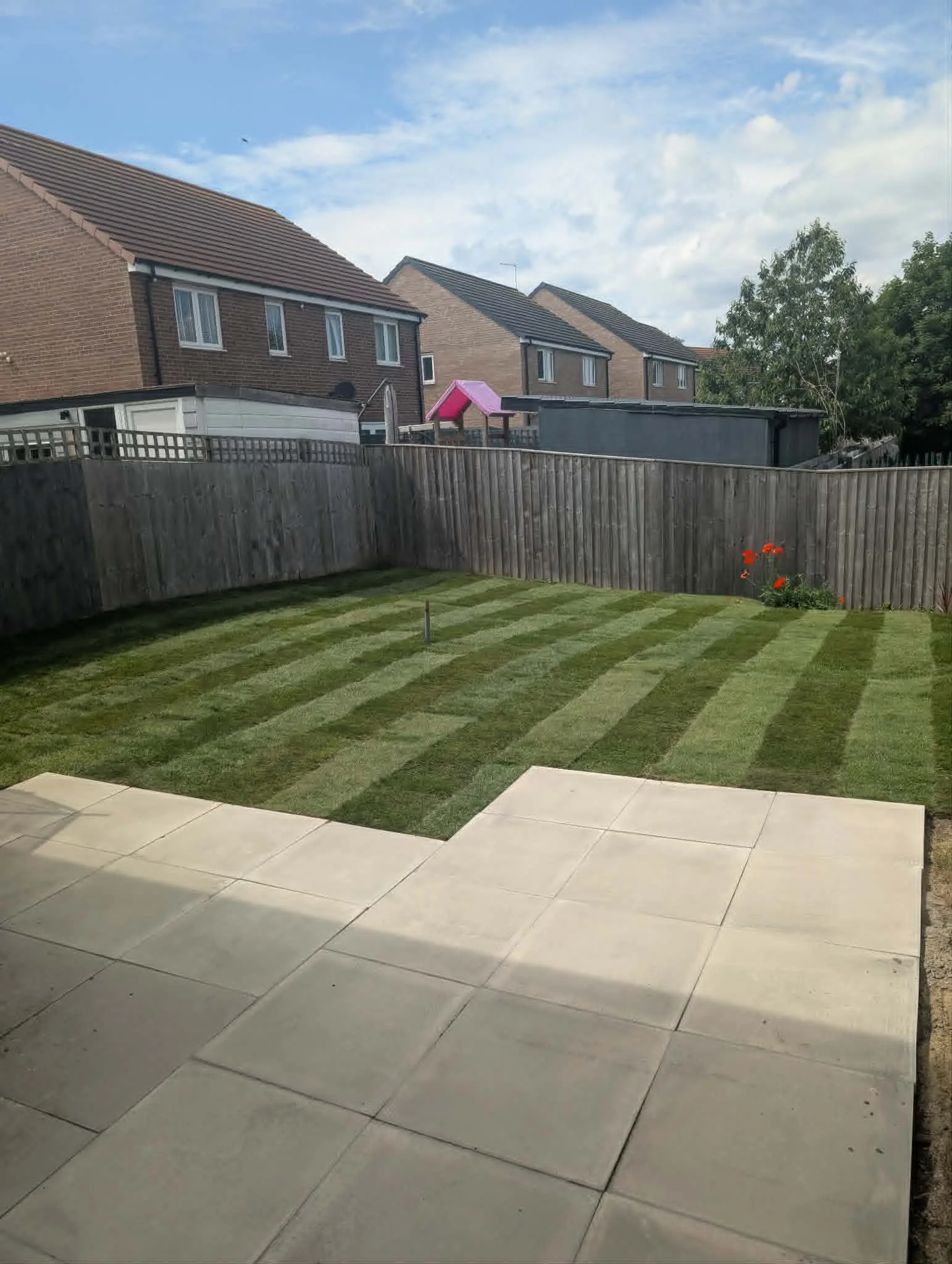 View of a backyard with a wooden fence, a well-maintained lawn with stripes, a small flower bed with red flowers, and neighboring houses with brick walls and tiled roofs under a partly cloudy sky.