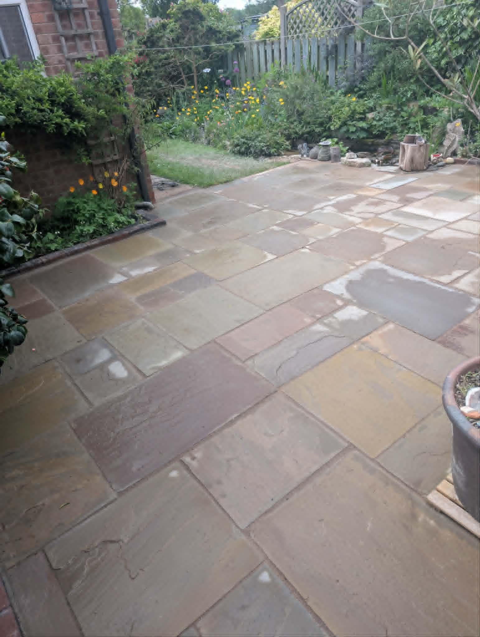 A backyard patio with stone paving, next to a grassy garden area with colorful flowers and a wooden fence.