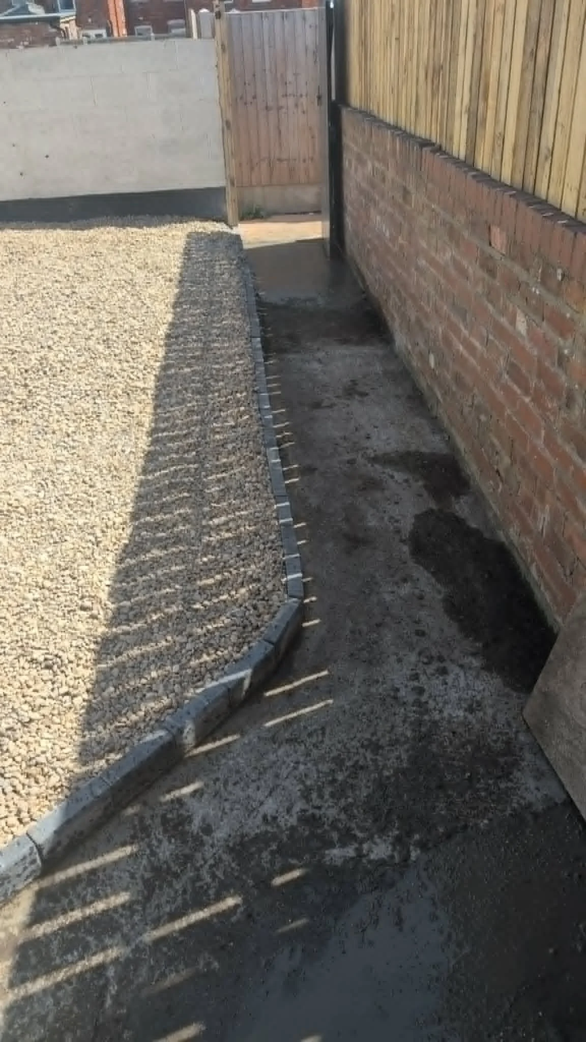 A narrow pathway lined with bricks, gravel, and concrete, bordered by a brick wall on the right and a wooden fence on the left, with shadows cast by the fence on the ground.