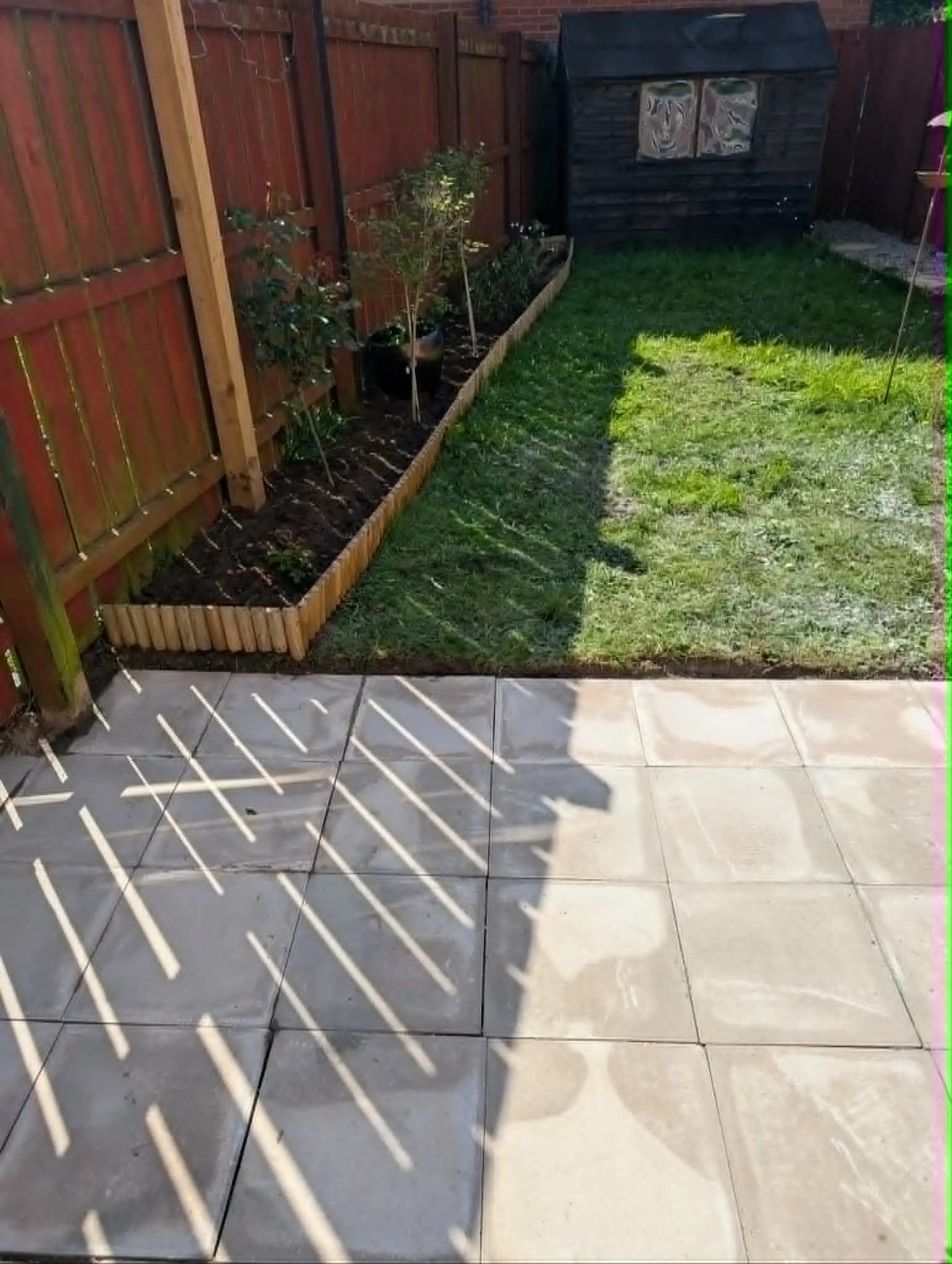 Backyard with tiled patio, grass, and a wooden raised garden bed with plants, enclosed by a wooden fence, and a small dark-colored shed in the corner.