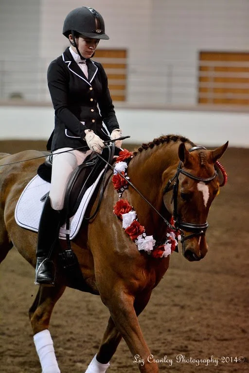 A female equestrian dressed in formal riding attire riding a victory lap on a decorated chestnut horse with a Reserve Champion garland around its neck in an indoor riding arena.