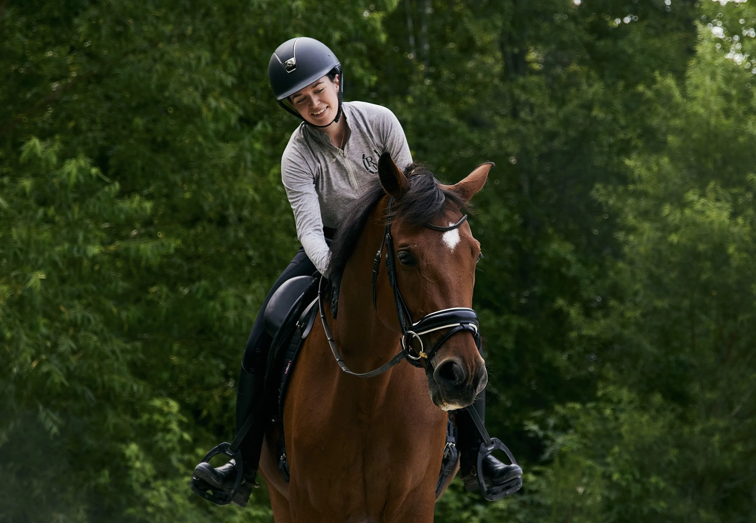 A young woman riding a brown horse in a green outdoor setting, wearing a riding helmet, a zip-up shirt, and riding gear.