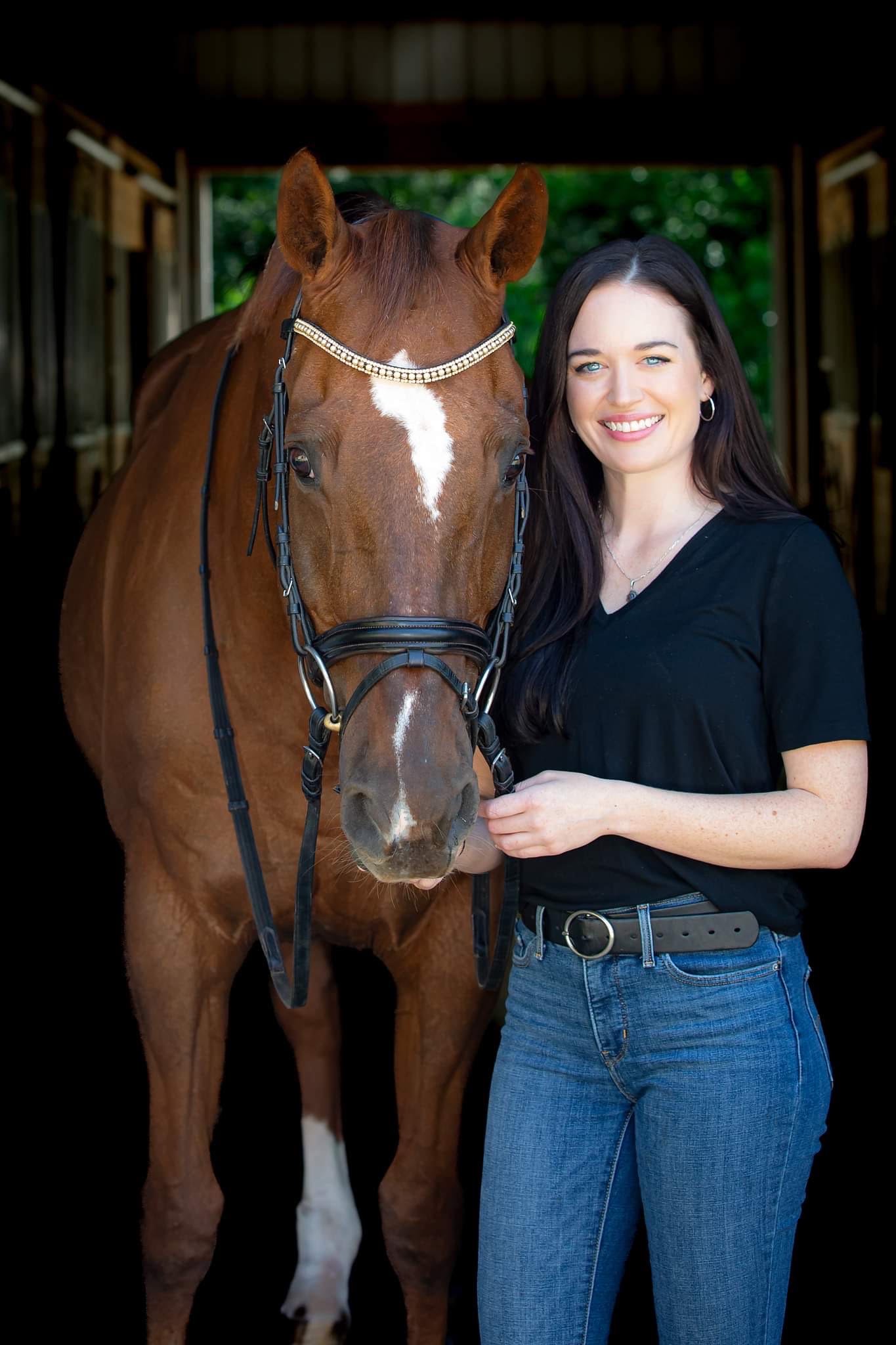 A woman with long dark hair, wearing a black shirt and blue jeans, standing next to a large brown horse in a stable with wooden walls and green trees in the background.