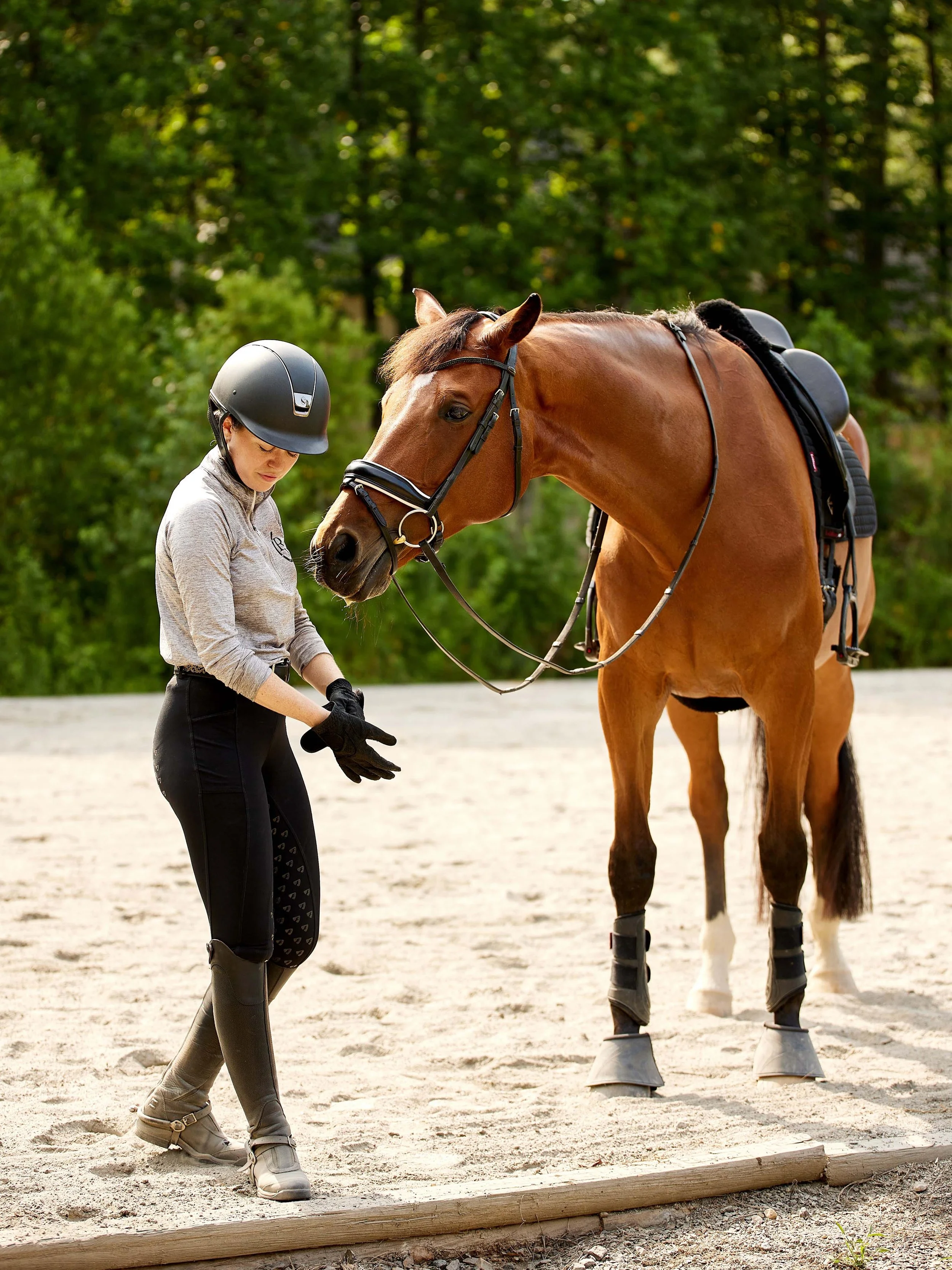 A woman wearing riding gear, including a helmet and tall riding boots, stands next to a brown horse with a black mane. The horse is fitted with riding tack and standing on a sandy arena with green trees in the background.