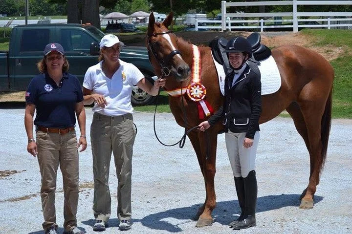 Three women and a horse with Reserve Champion High Point Awards, standing outdoors on a gravel surface with cars and a white fence in the background.
