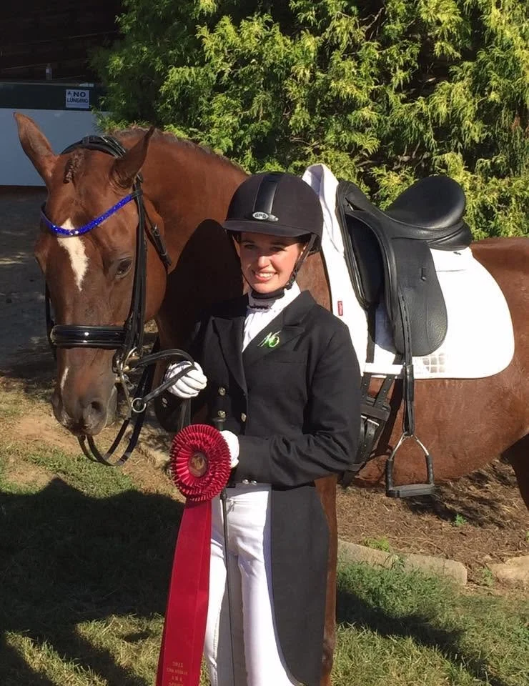 A smiling woman in equestrian attire holding a National Top Ten ribbon standing next to a brown horse with a white star-shaped mark on its face. The horse is equipped with a bridle and saddle.