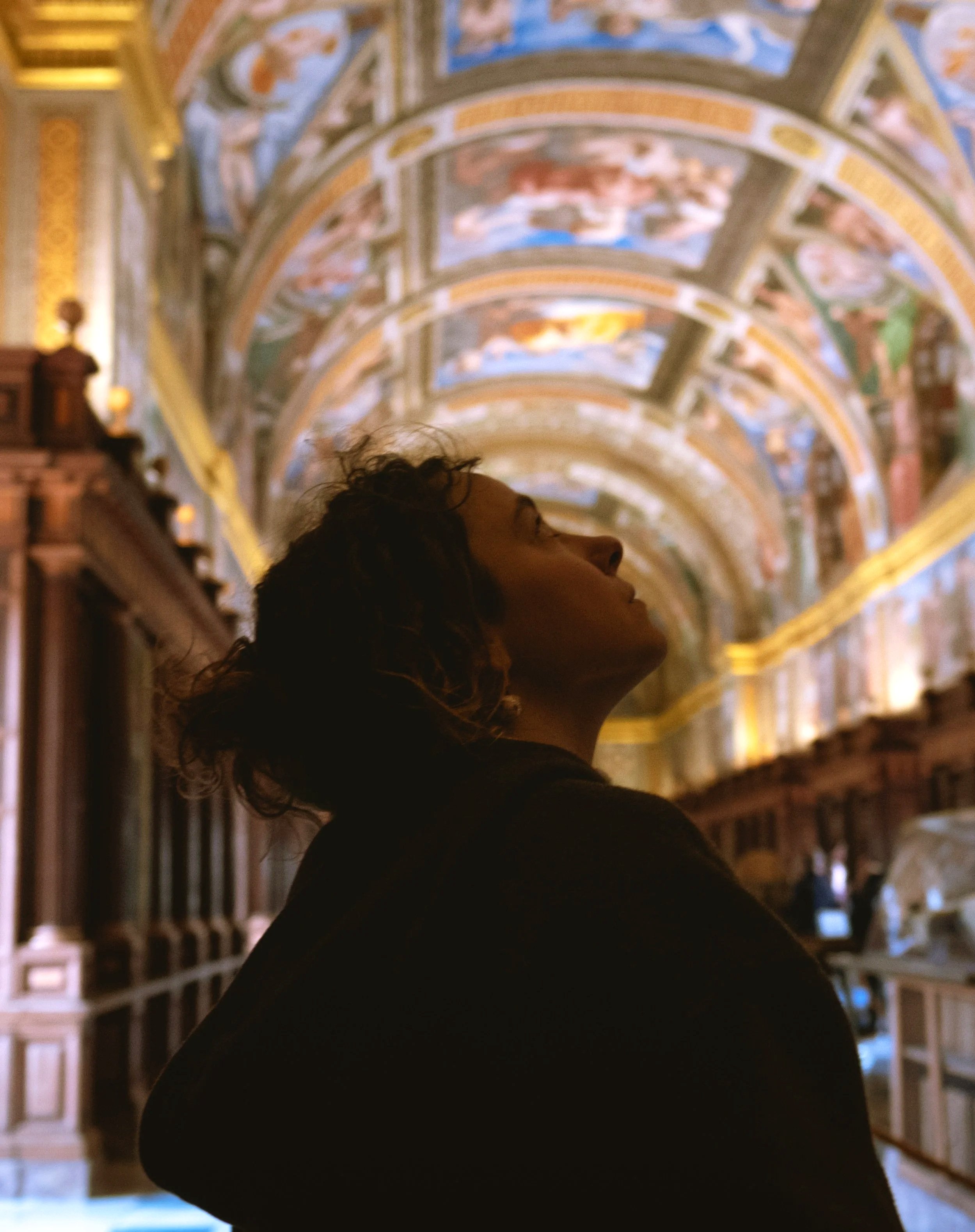 A person with dark curly hair is standing in front of a large, ornate, painted ceiling in a library or museum with a high, arched ceiling decorated with detailed frescoes and gold accents.