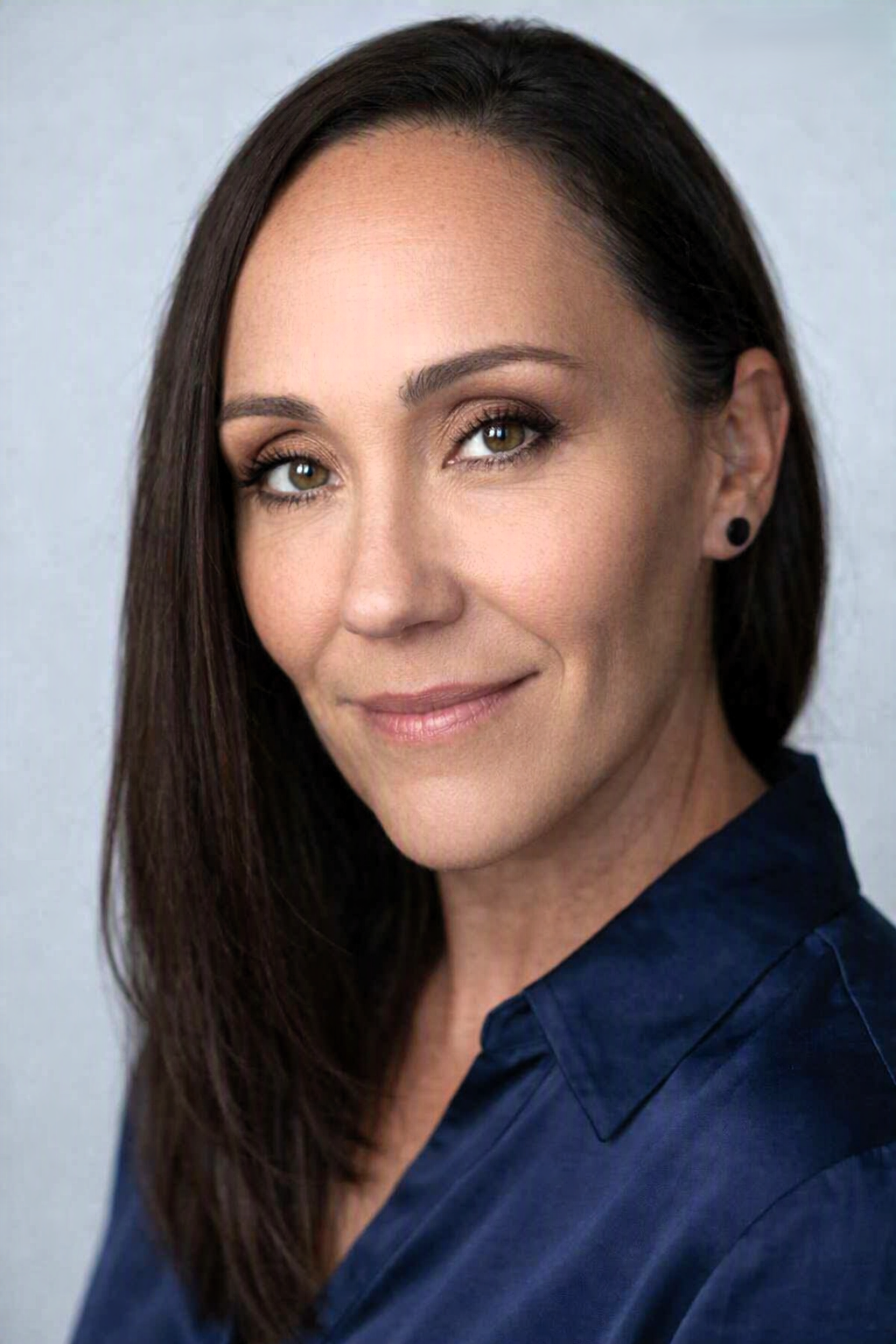 Close-up portrait of a woman with long dark hair, wearing makeup, a dark blue shirt, and small black earrings, smiling at the camera against a plain light background.