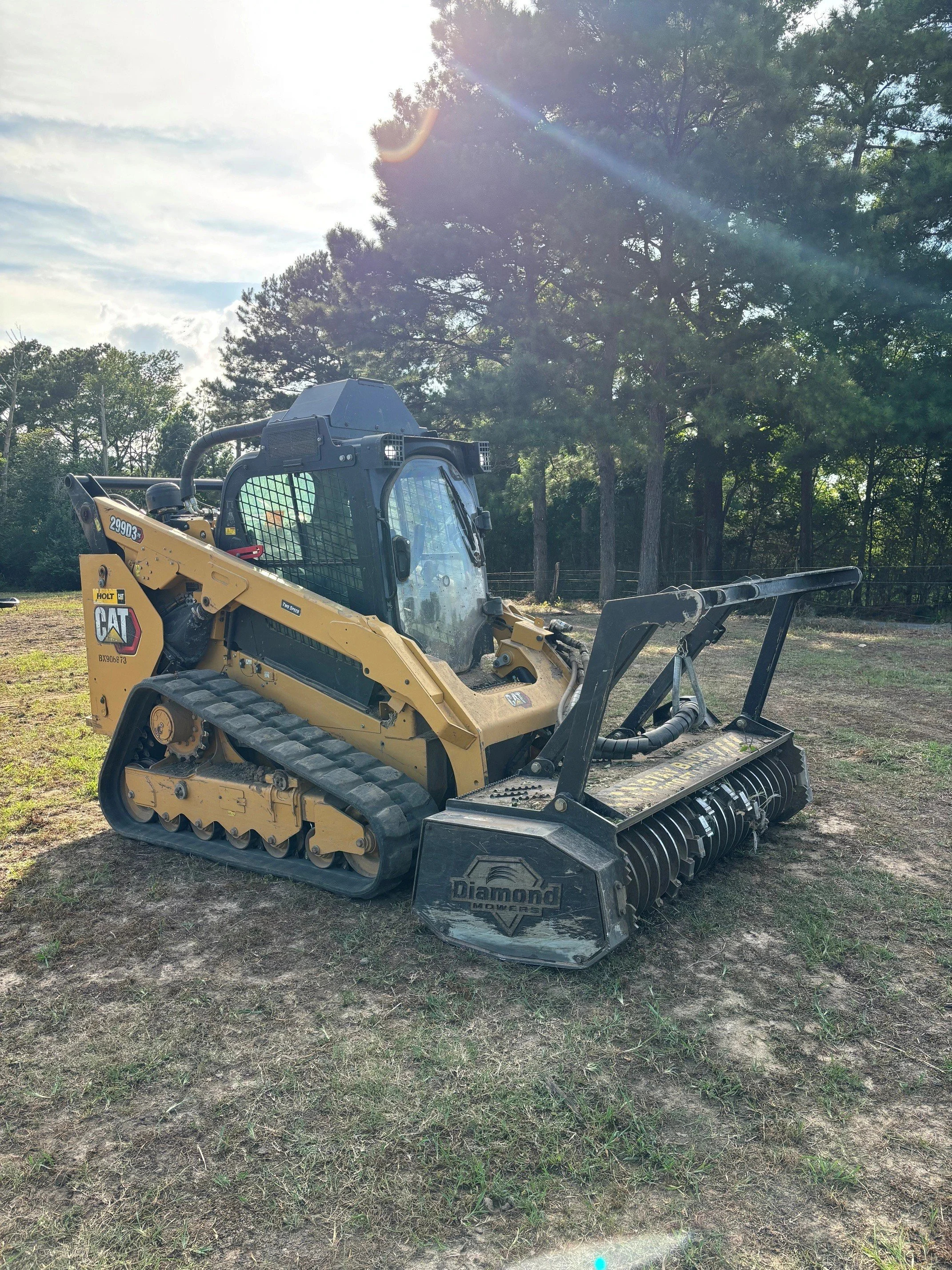 A yellow Caterpillar compact construction excavator with a black cab parked on a grassy patch, surrounded by tall trees under a partly cloudy sky with sunlight and lens flare.