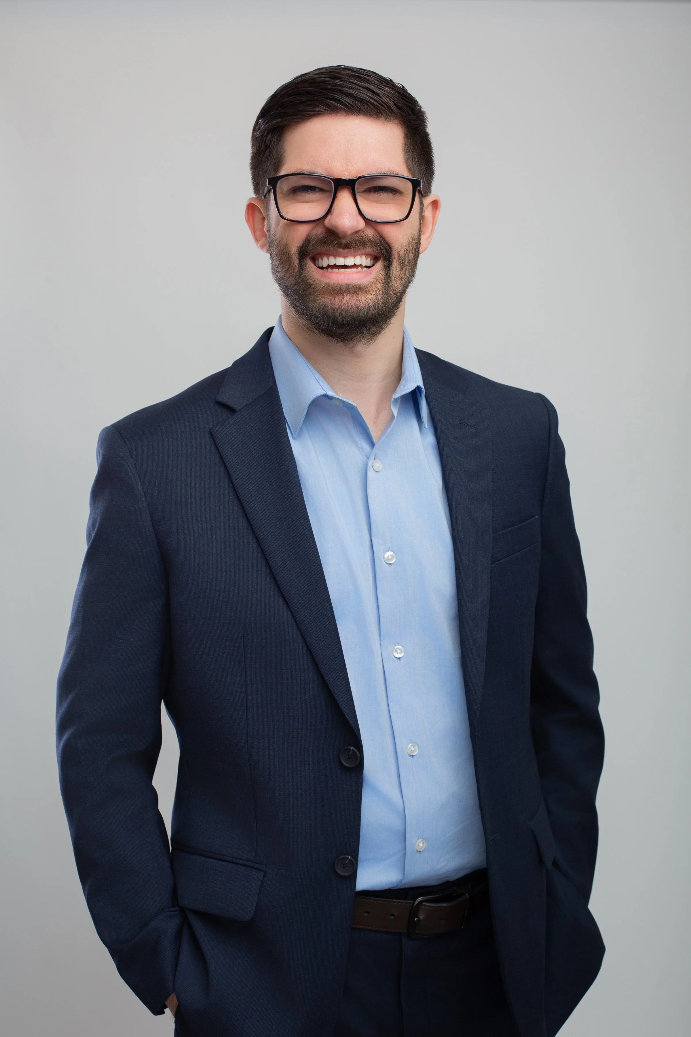 A man with dark hair and beard, wearing glasses, a navy suit, and a light blue shirt, smiling against a plain gray background.