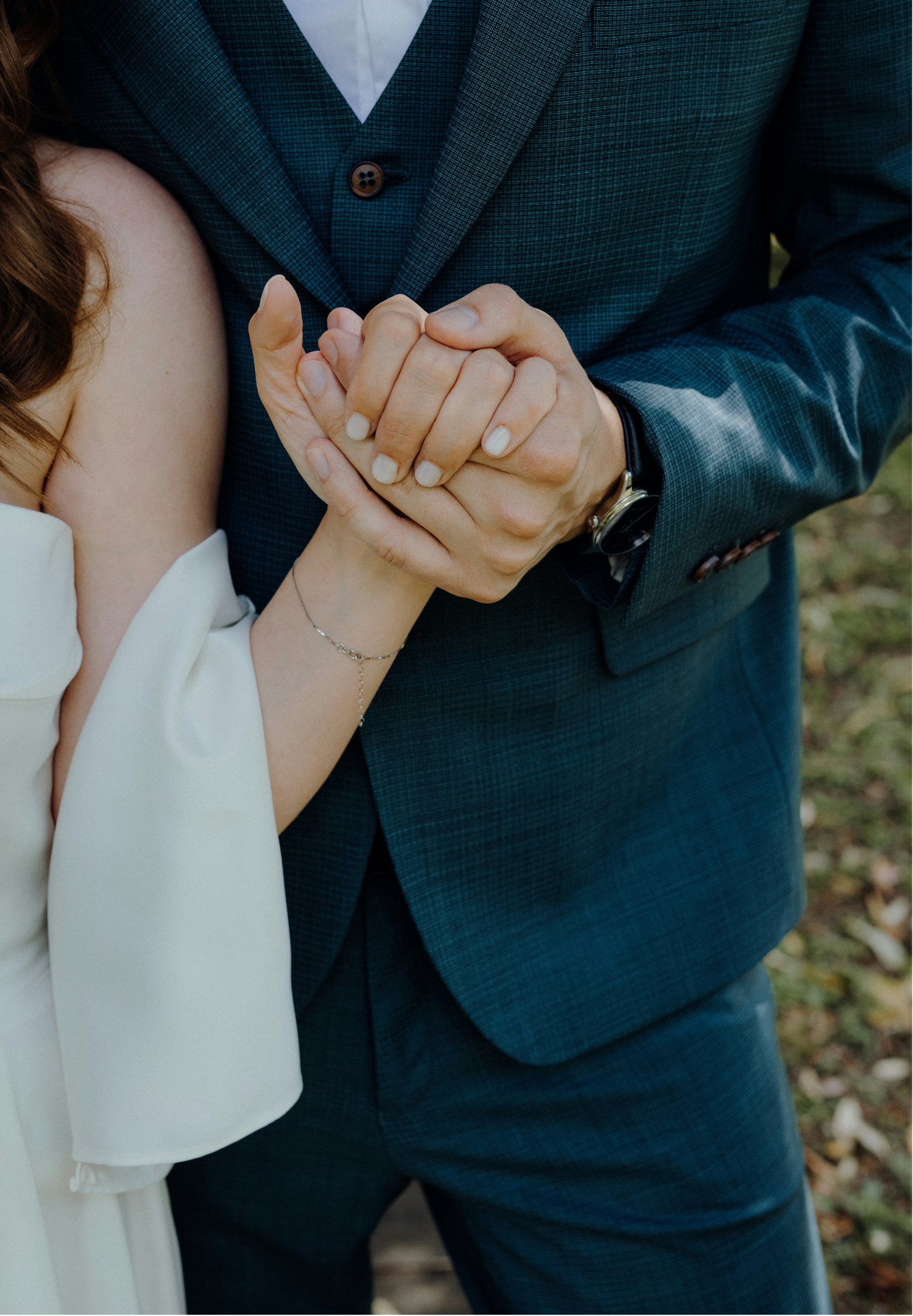Close-up of a man and a woman holding hands, with the man wearing a dark checkered blazer and the woman wearing a white dress, outdoors with leaves on the ground.