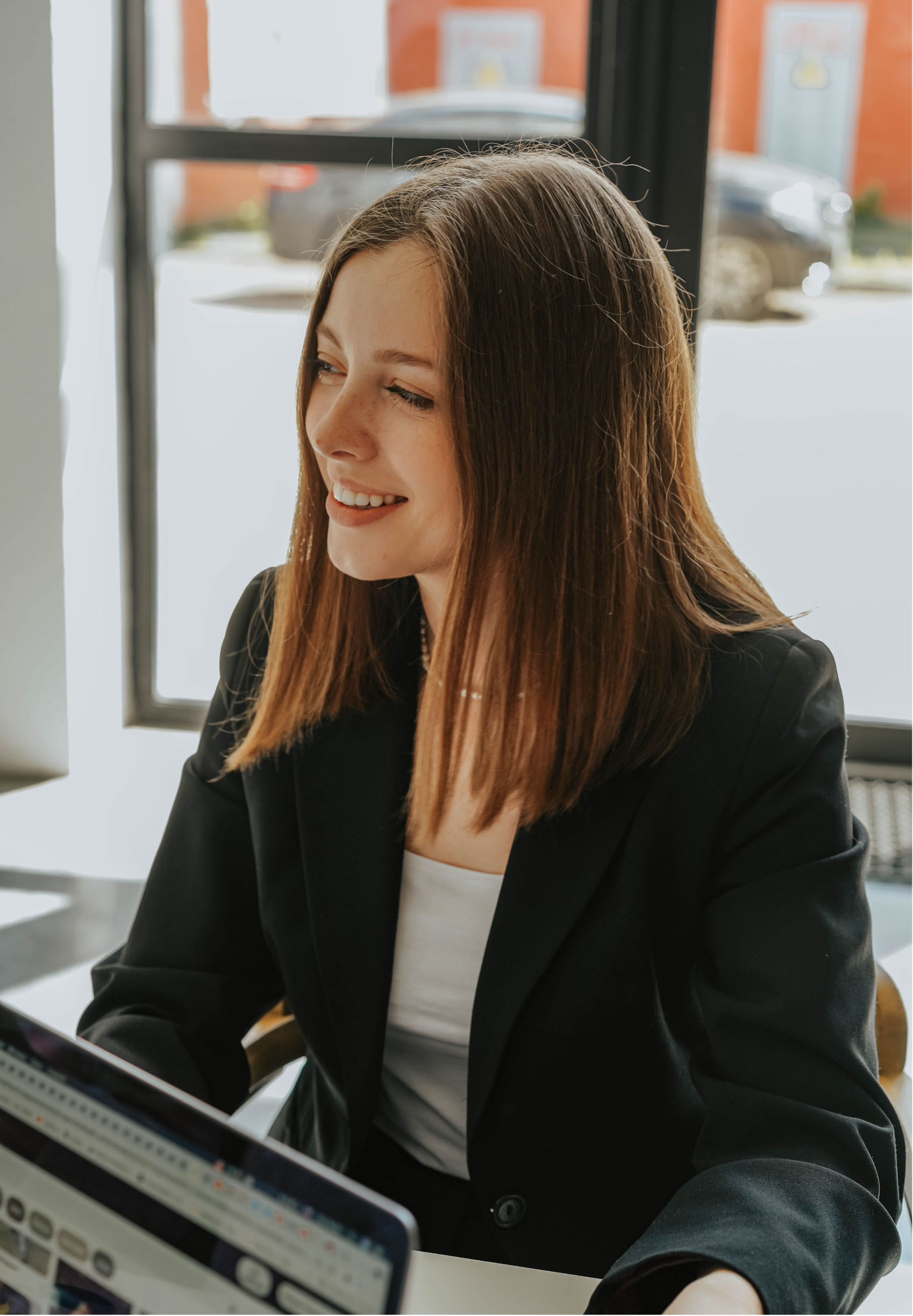 A smiling woman with long brown hair, wearing a black blazer and a white top, working on a laptop in a bright office with large windows.
