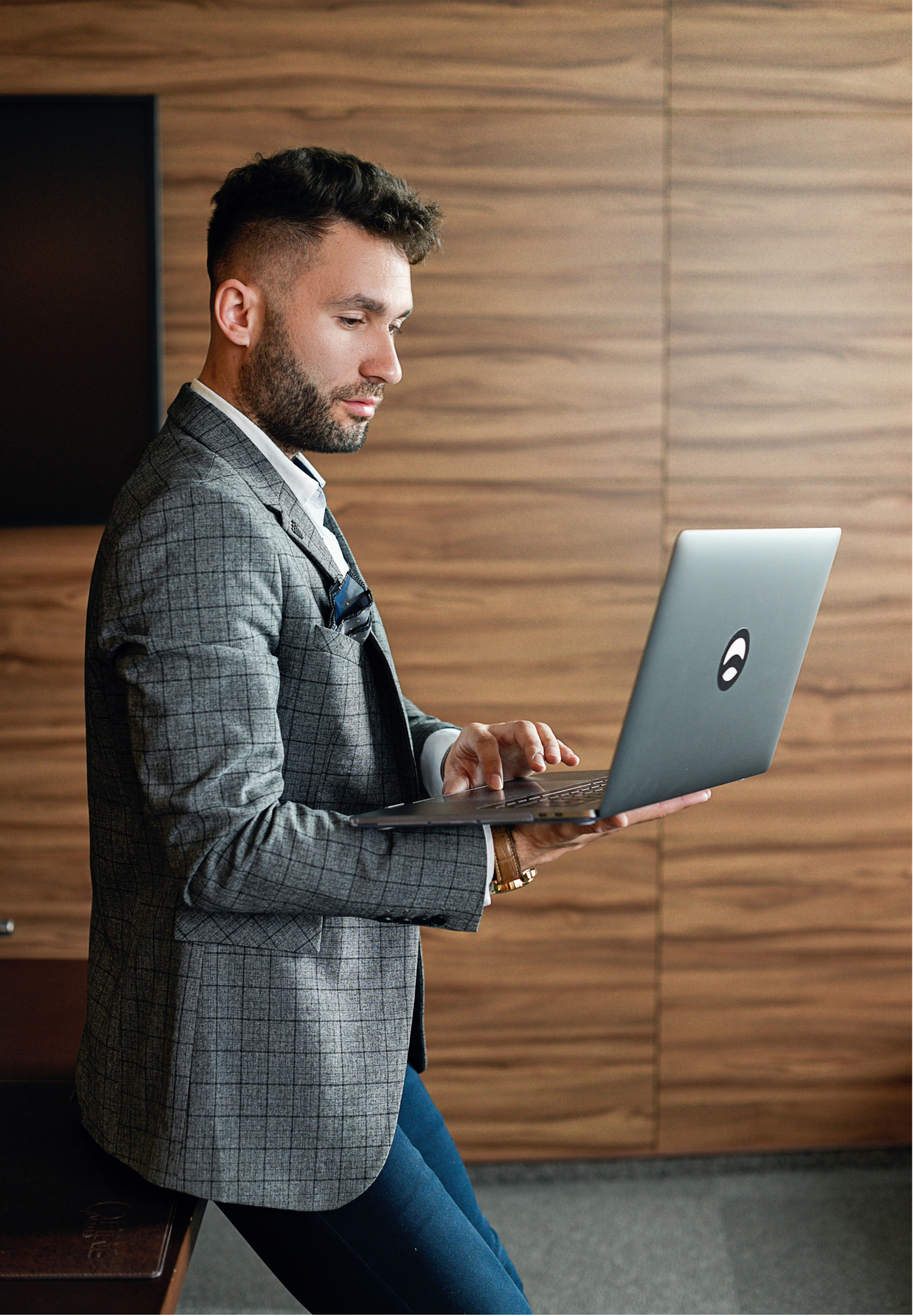 A man in a gray checkered blazer and dark blue jeans sitting in a room with wooden wall panels, looking at his open laptop.