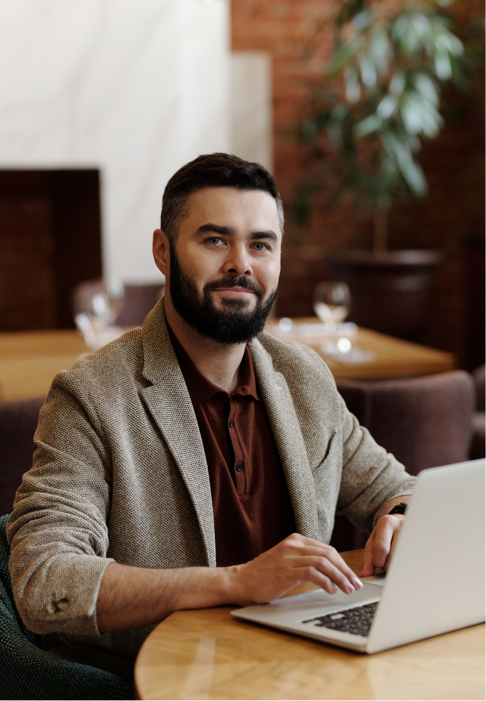 A man with a beard and short dark hair sitting at a wooden table, using a laptop with a white body and black keyboard. He is wearing a brown shirt and a beige checkered blazer. The background features a brick wall, some greenery, and out-of-focus table settings with glasses.