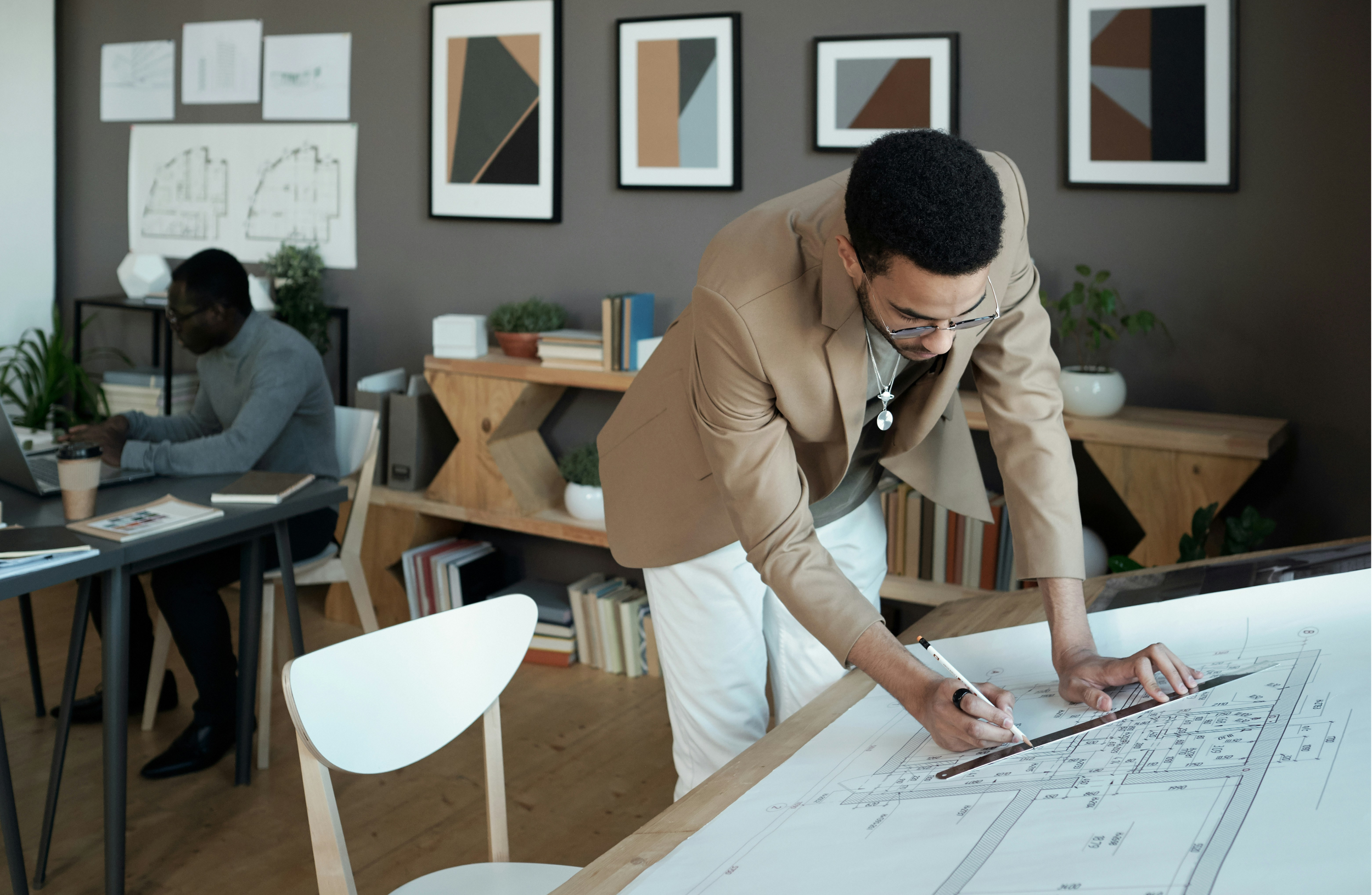 Two men working on architectural blueprints in an office with framed artwork and books on the shelves.