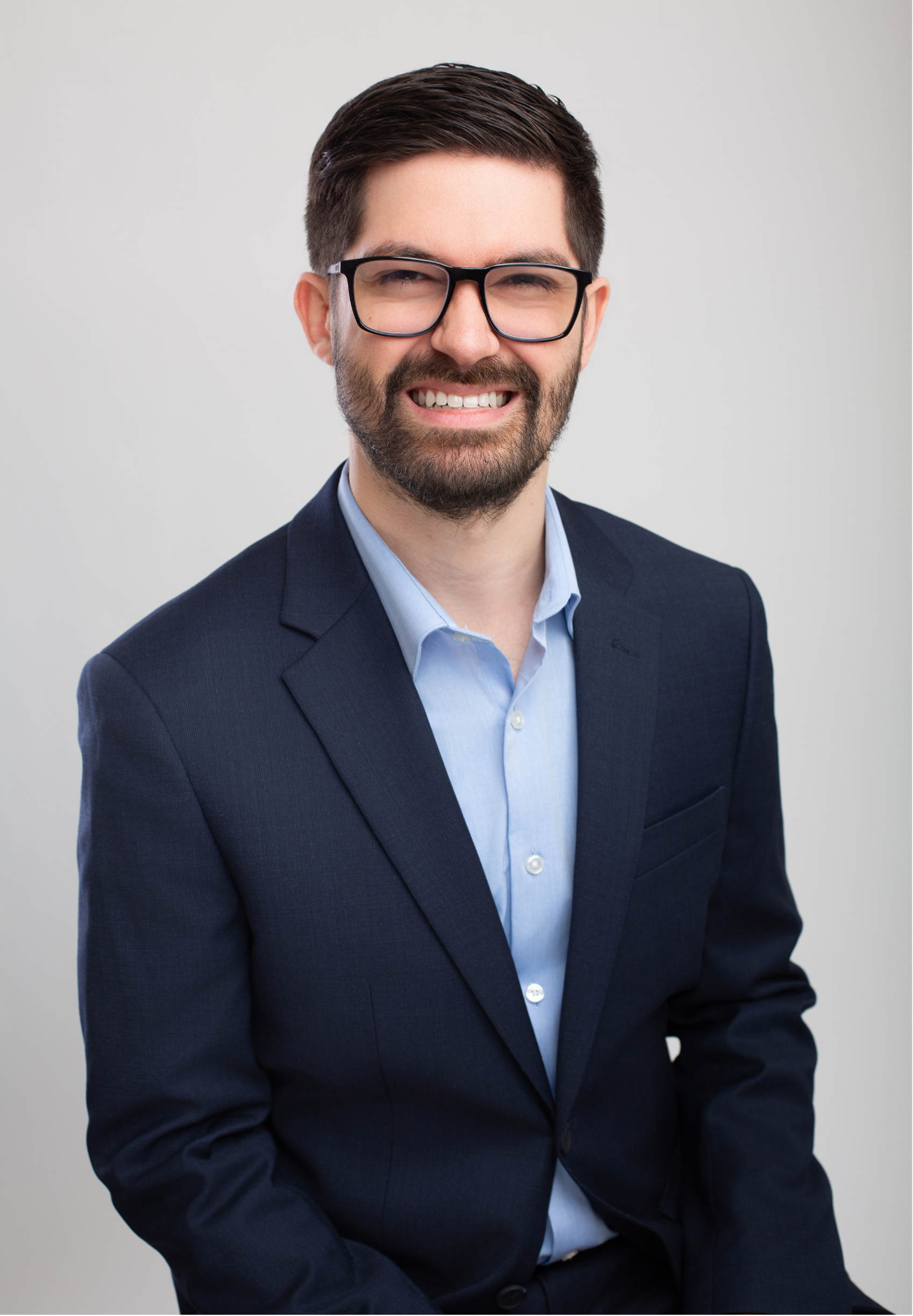 Dr. Joe Rustum, licensed clinical psychologist and executive coach in Nashville, TN, with dark hair, glasses, and a beard wearing a navy blazer and light blue shirt smiling against a gray background.