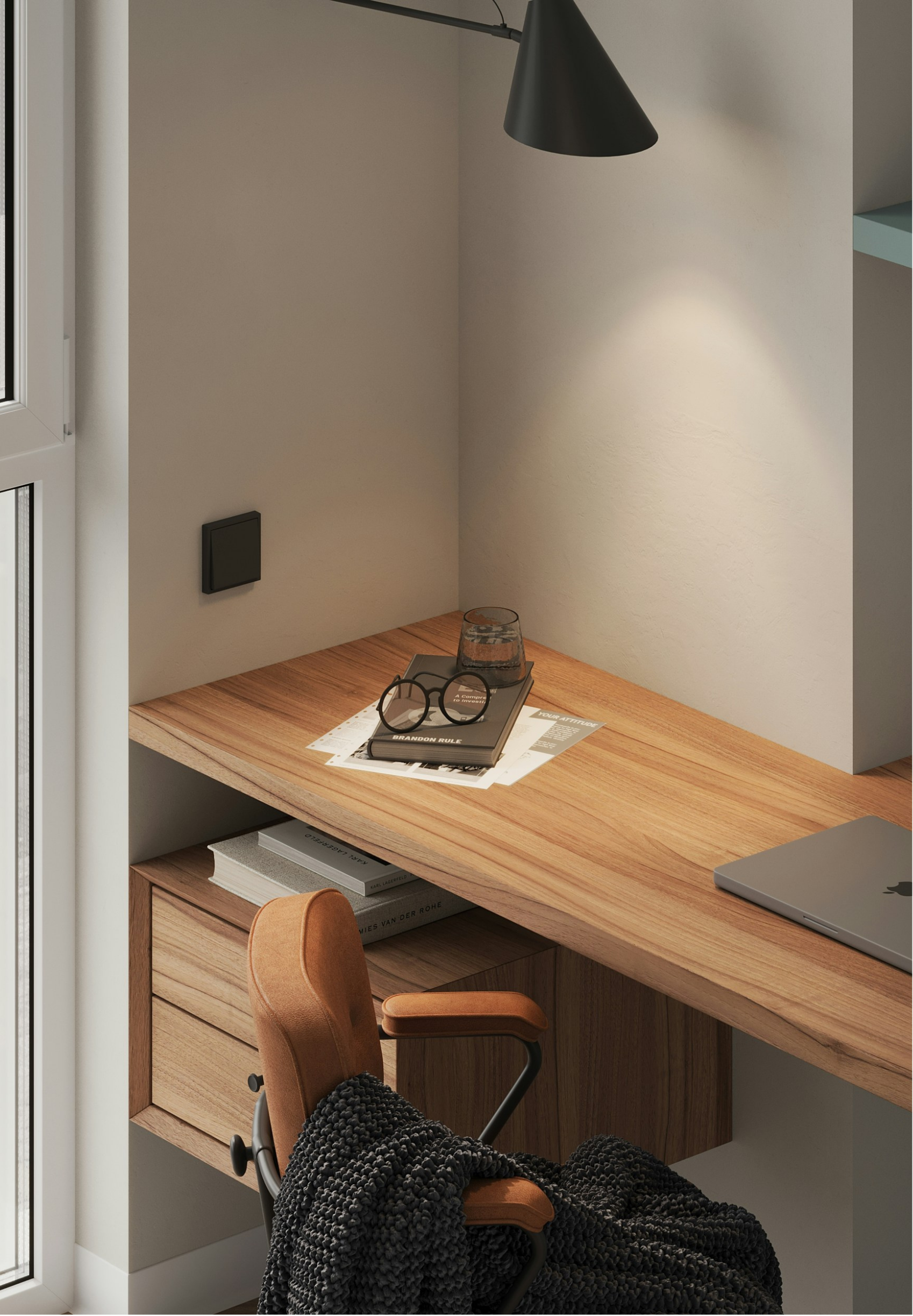 Corner of a wooden desk with a chair, a laptop, and a black textured blanket. On the desk, two stacked books, a glass of water, glasses, and papers. Wall behind the desk with a light switch, a small rectangular black device, and a wall lamp. A window is visible on the left side.