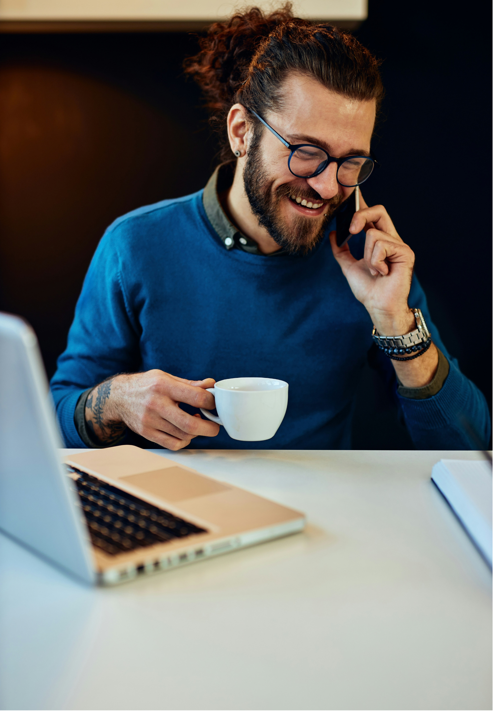 A man with glasses and a beard, wearing a blue sweater, smiling while talking on a phone, holding a white coffee mug, sitting at a desk with a laptop and notebook.