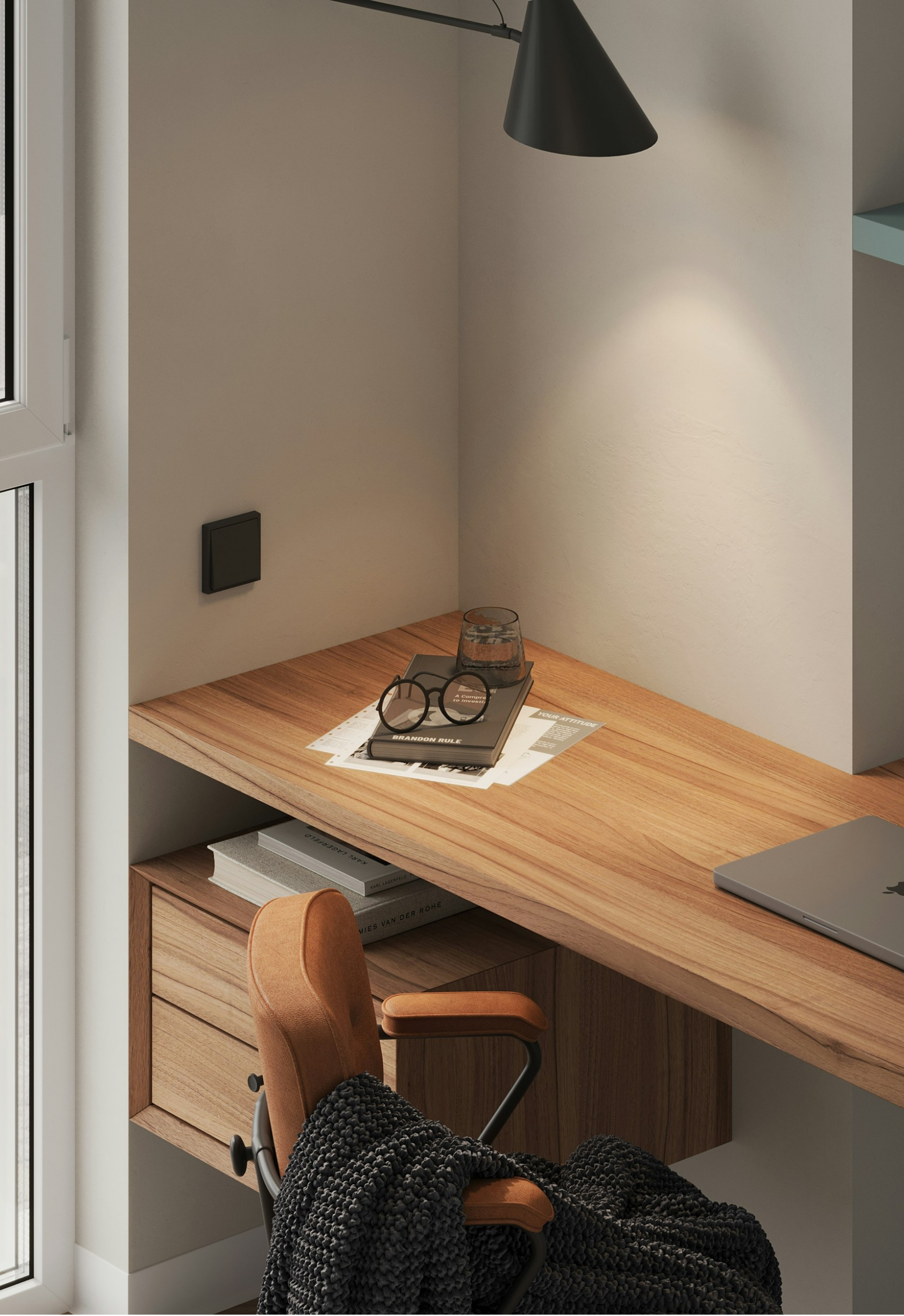 Corner of a home office with a wooden desk, a brown office chair, a black wall lamp, a pair of glasses, and some books on the shelf underneath.