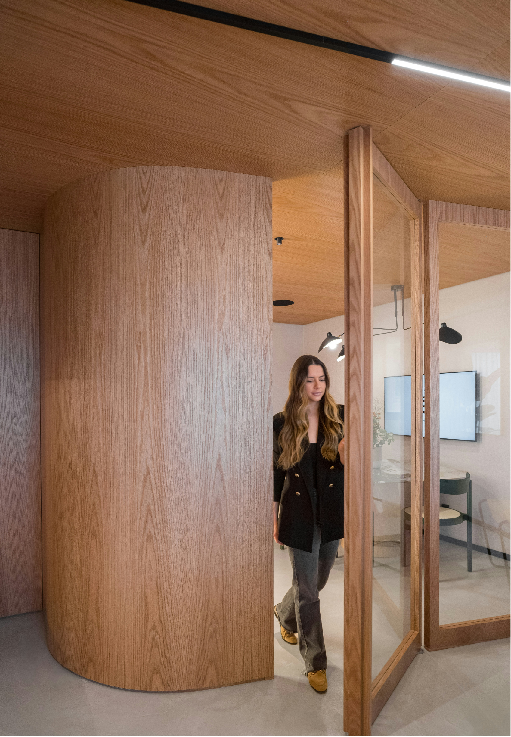 Woman with long wavy hair wearing a black blazer and gray pants walking through a wooden partition of a modern interior space.