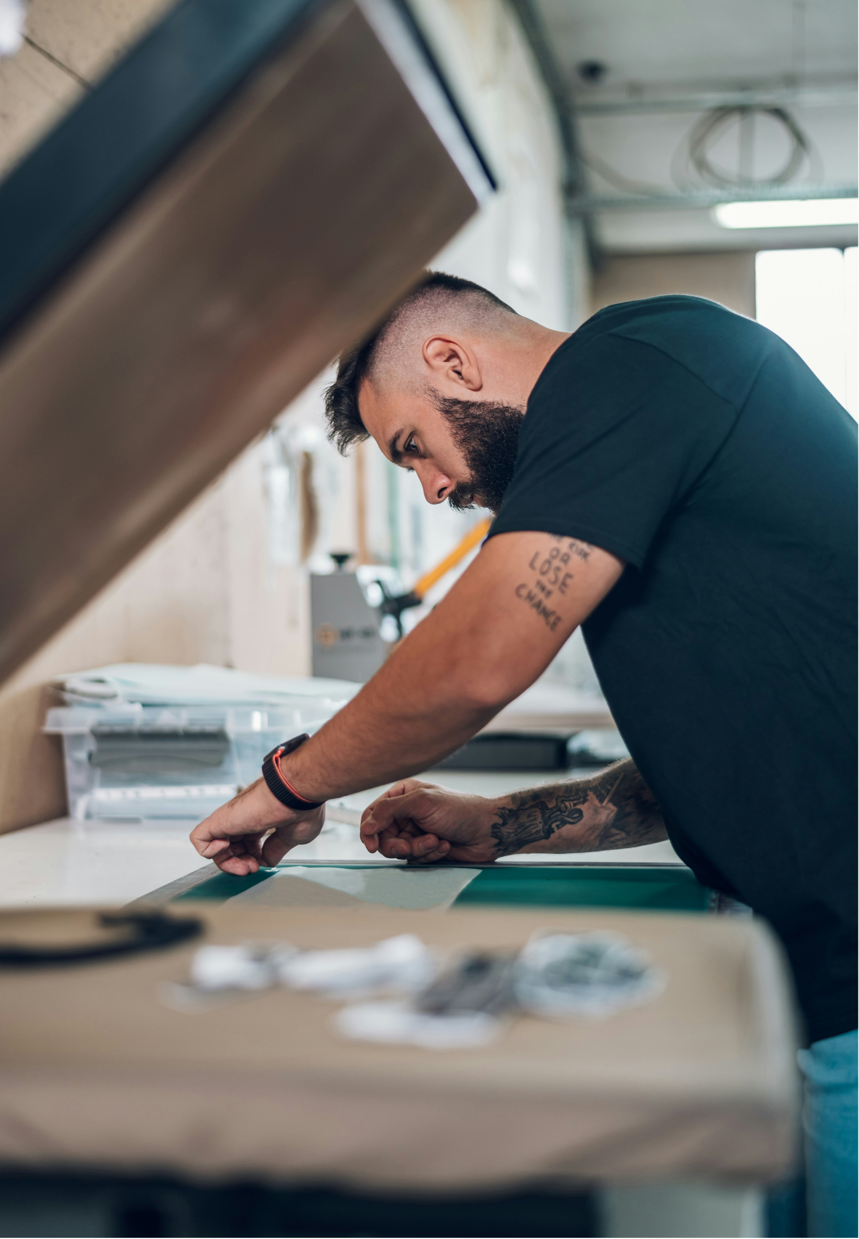 A man with a beard and tattoos on his arm working at a workstation, leaning over a table with papers or materials, in a well-lit room with industrial ceiling pipes, window, and natural light.