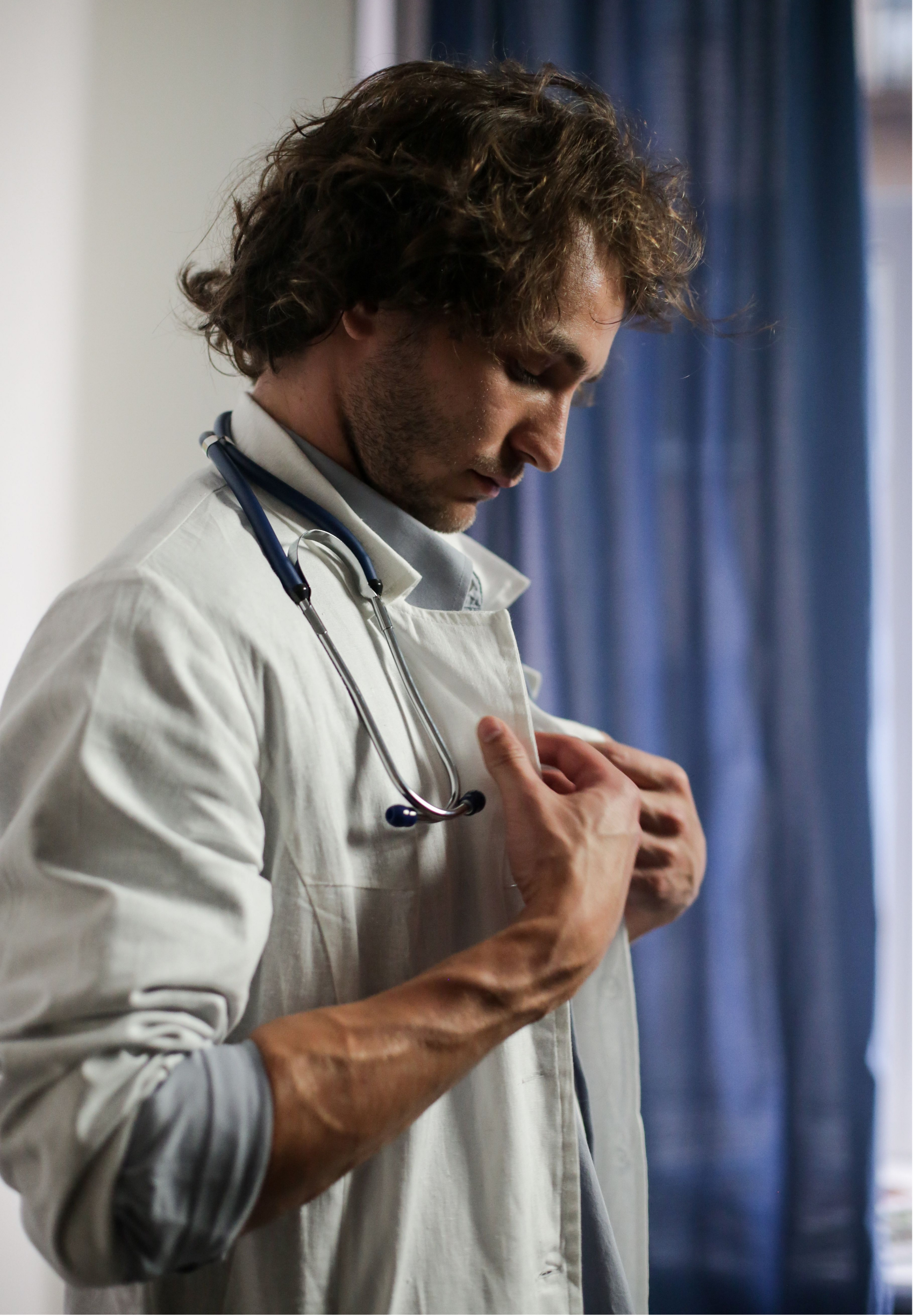 A male healthcare worker with curly brown hair, wearing a white coat and a stethoscope around his neck, is standing with his head bowed, and his hands placed on his chest.