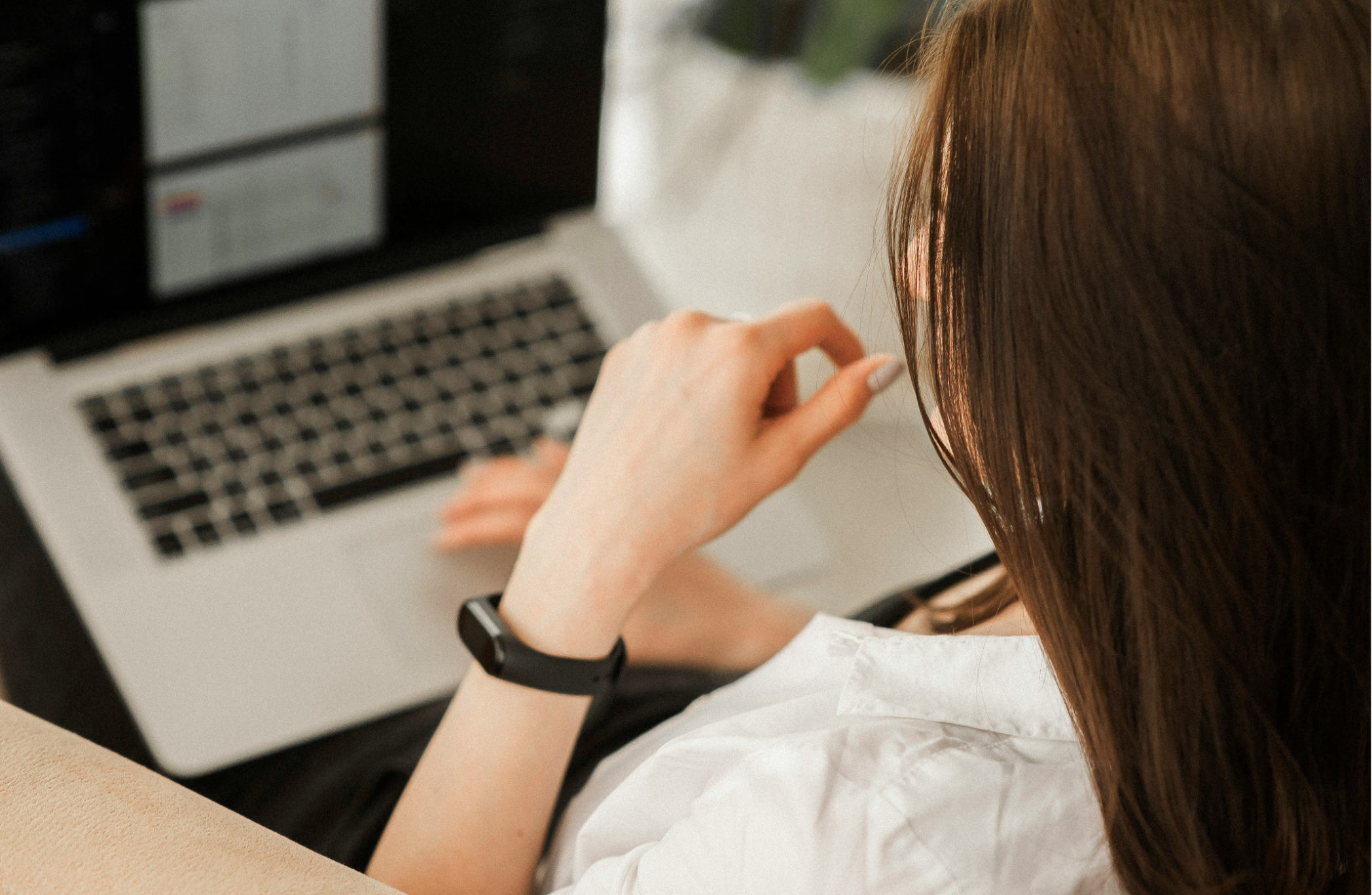 Person with long brown hair wearing a white shirt, black smartwatch, and a black fitness tracker, sitting in front of a laptop with the screen slightly blurred.