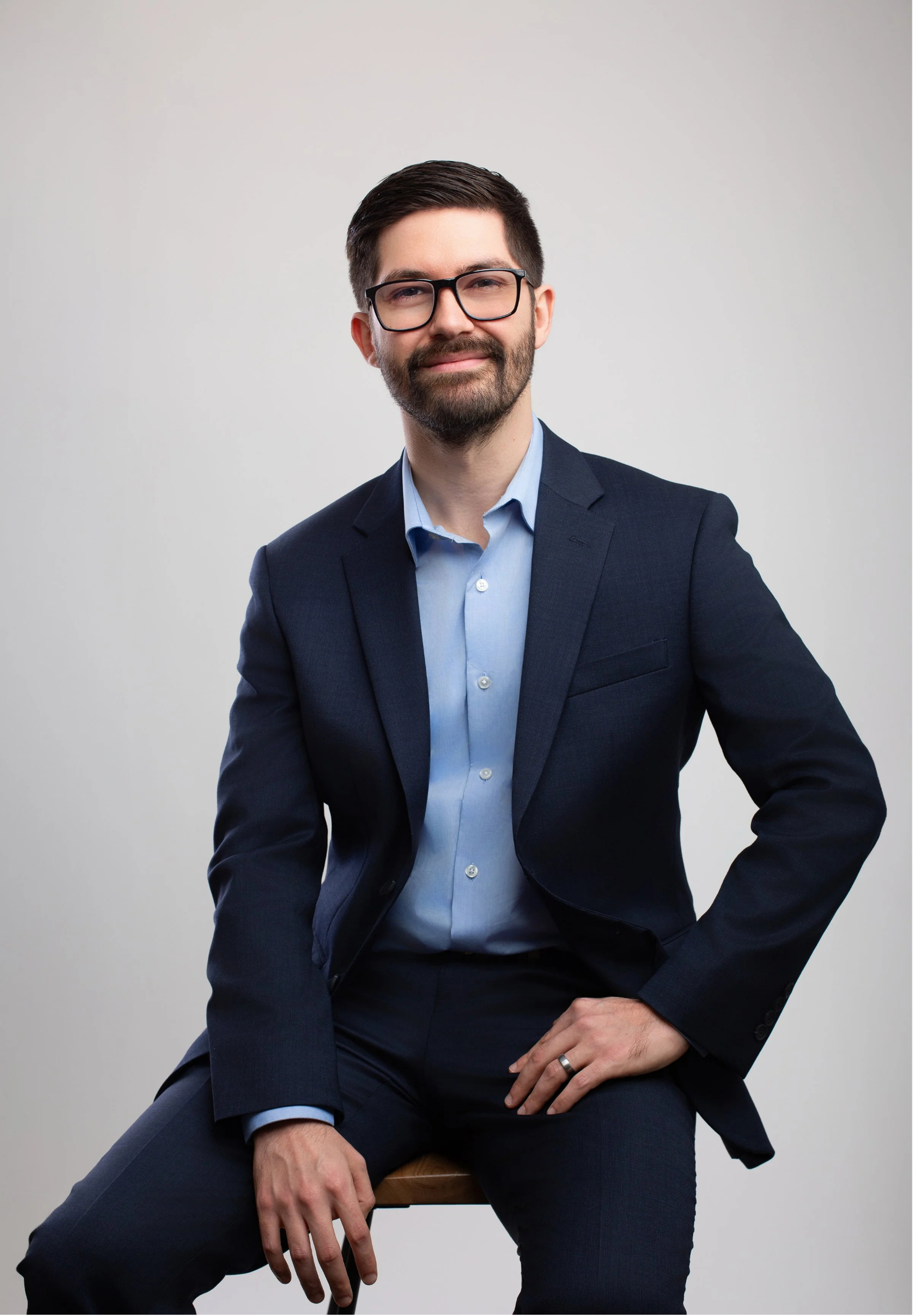 Dr. Joe Rustum, licensed clinical psychologist and executive coach in Nashville, TN, with glasses and a beard wearing a business suit, sitting on a stool against a plain gray background.