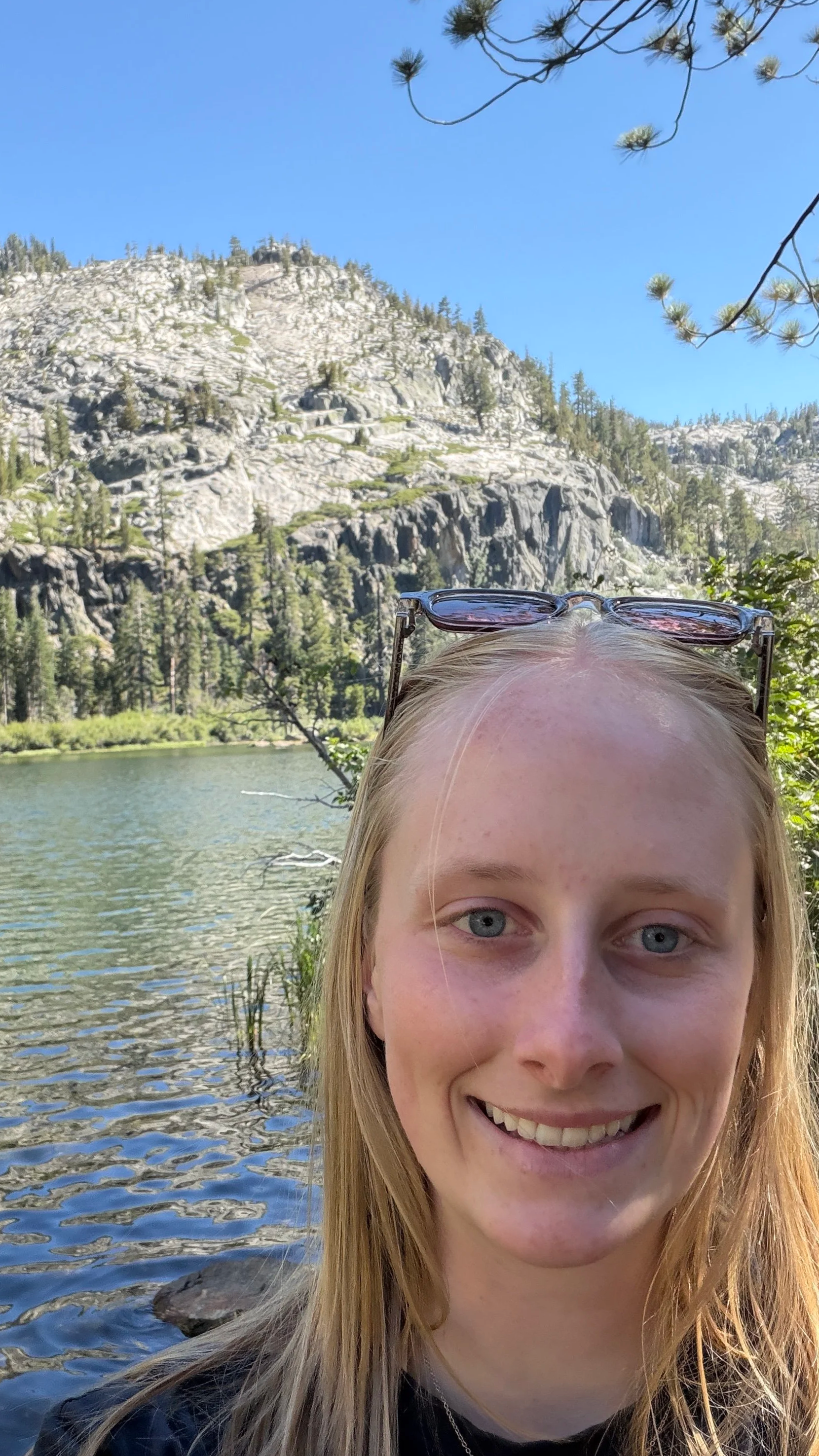 selfie near a lake with a mountain and pine trees in the background on a sunny day.