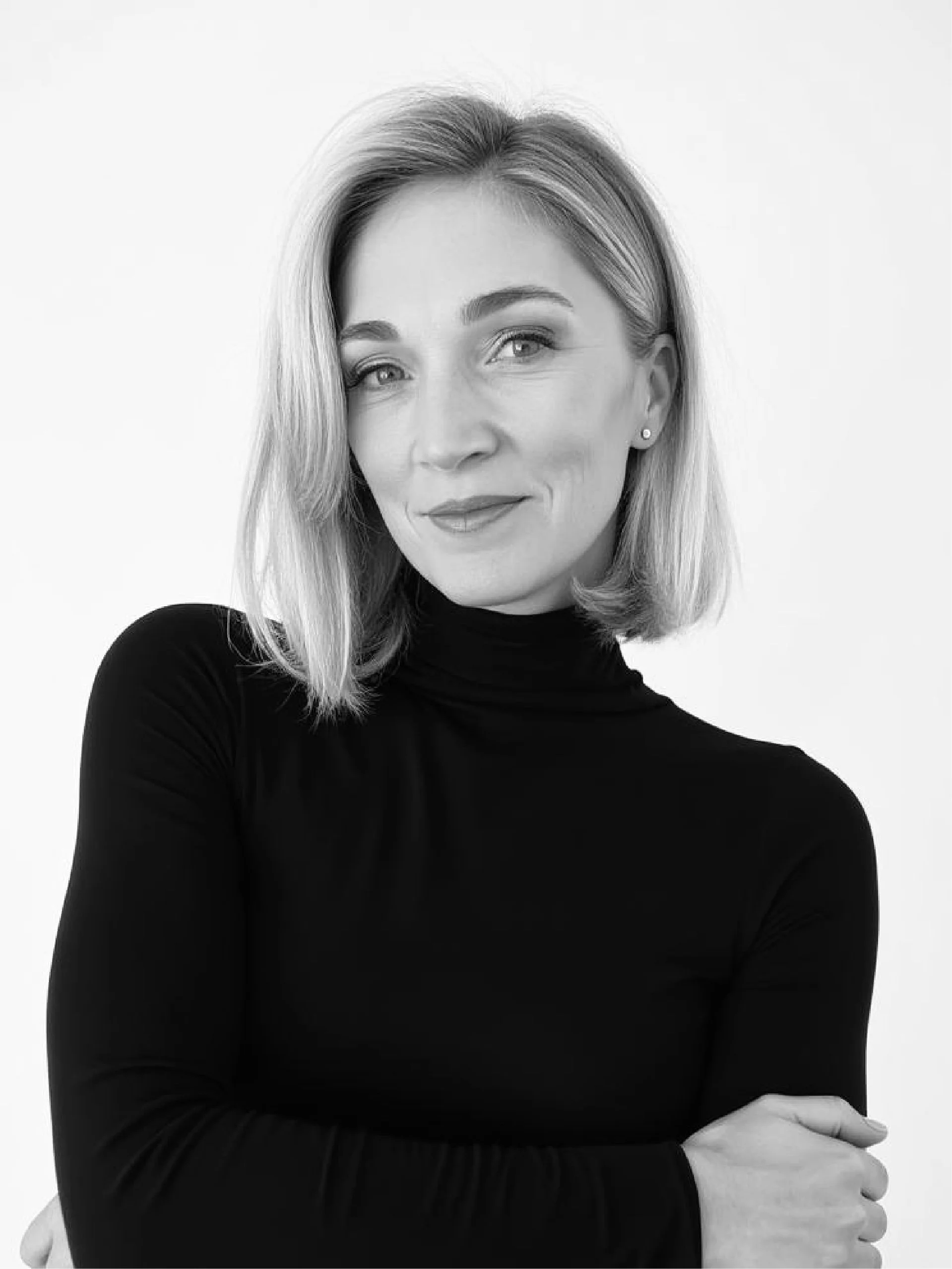 Black and white portrait of a woman with shoulder-length hair, wearing a long-sleeved black top, smiling softly with her arms crossed against a plain light background.