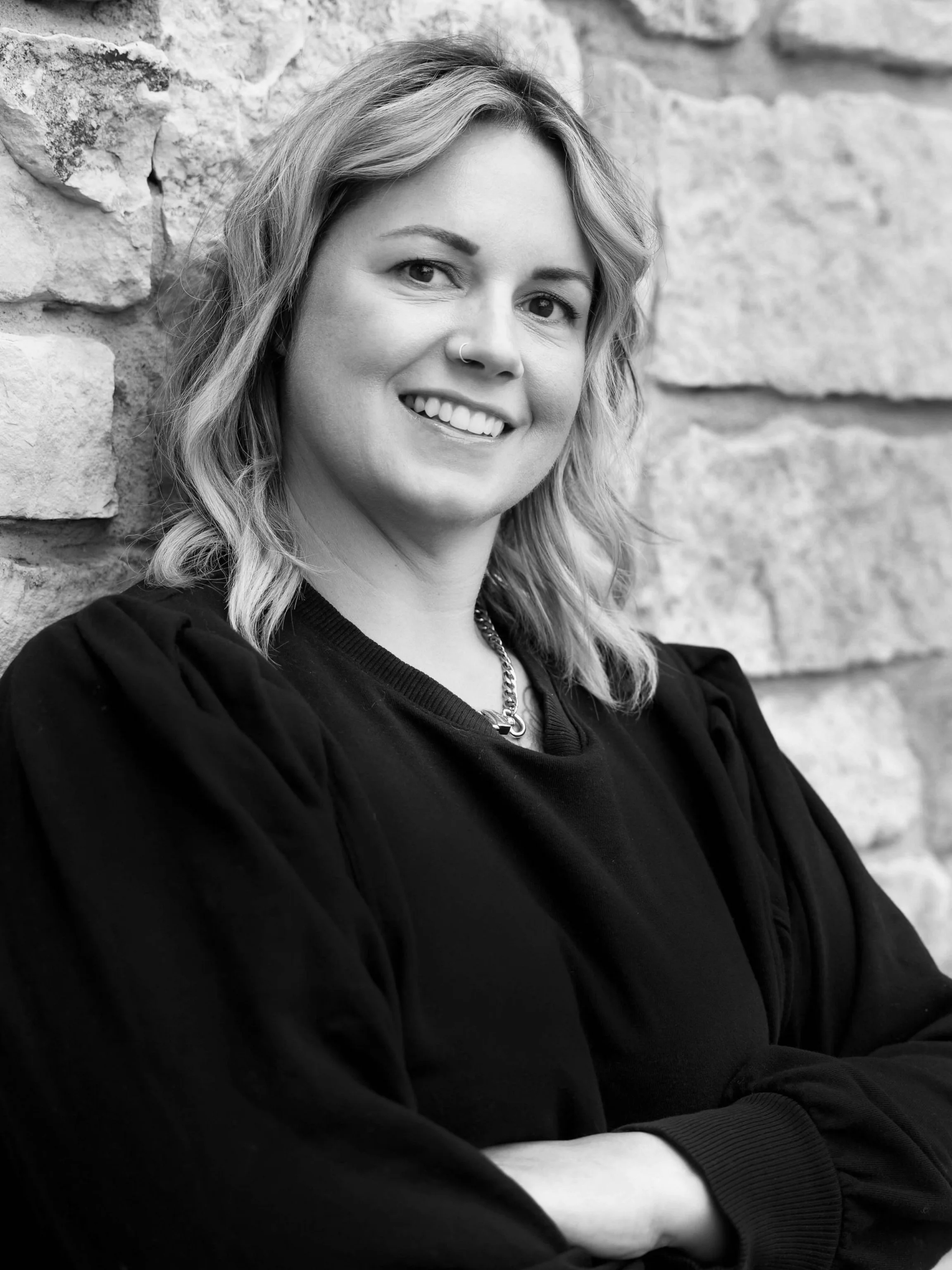 Black and white photograph of a woman with shoulder-length wavy hair, smiling, wearing a nose ring and a chain necklace, standing against a stone wall.
