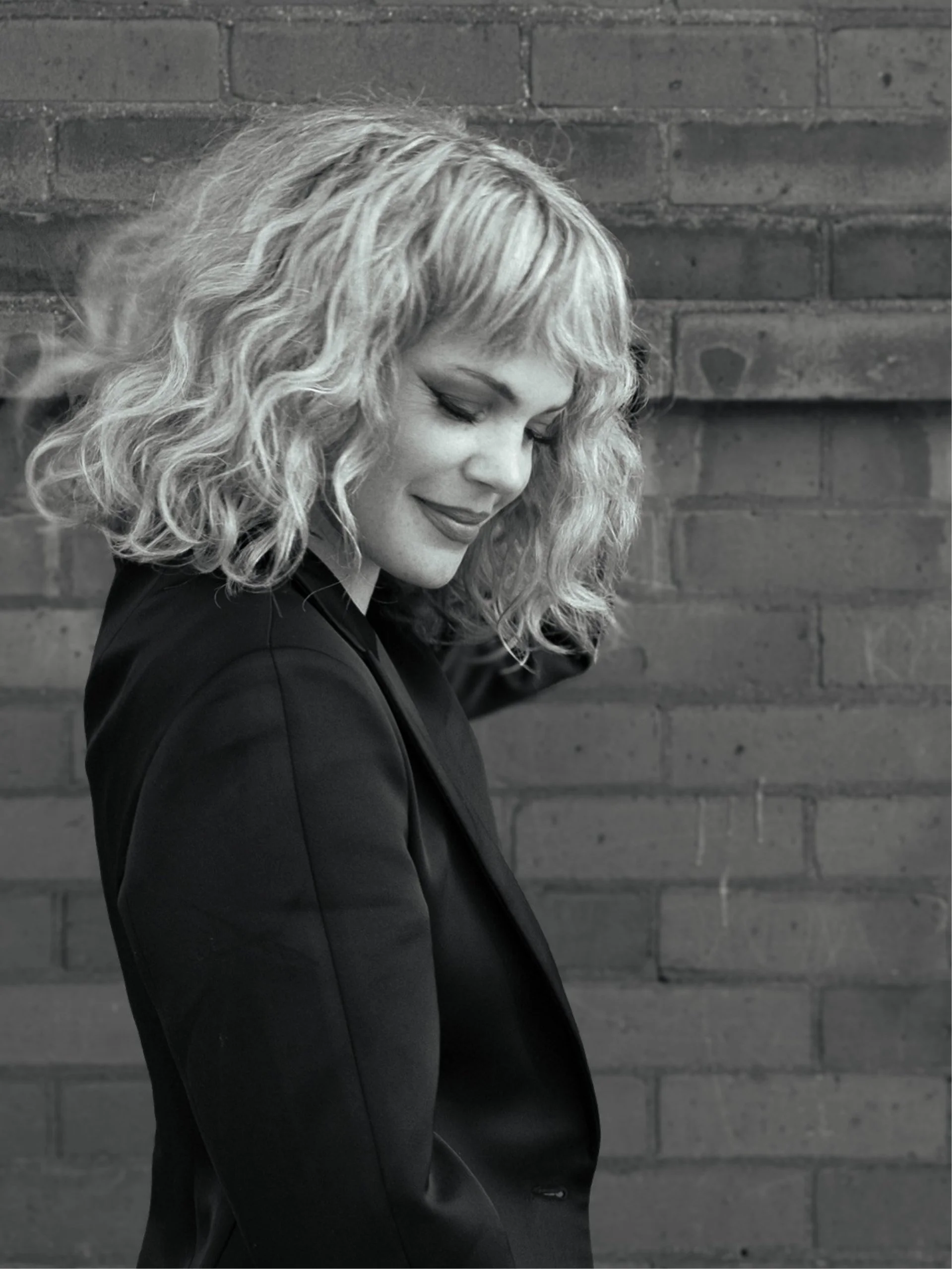 A woman with curly, shoulder-length blonde hair, smiling with her eyes closed, wearing a black blazer, standing against a brick wall.
