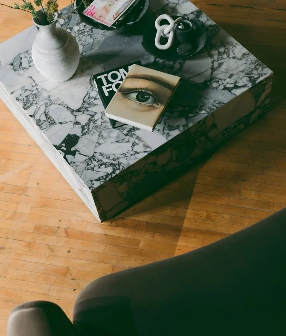 A marble-topped coffee table with a white vase of flowers, a book with a close-up of an eye on the cover, a black and white magazine, a black dish with white decorative knots, and some papers on a hardwood floor.