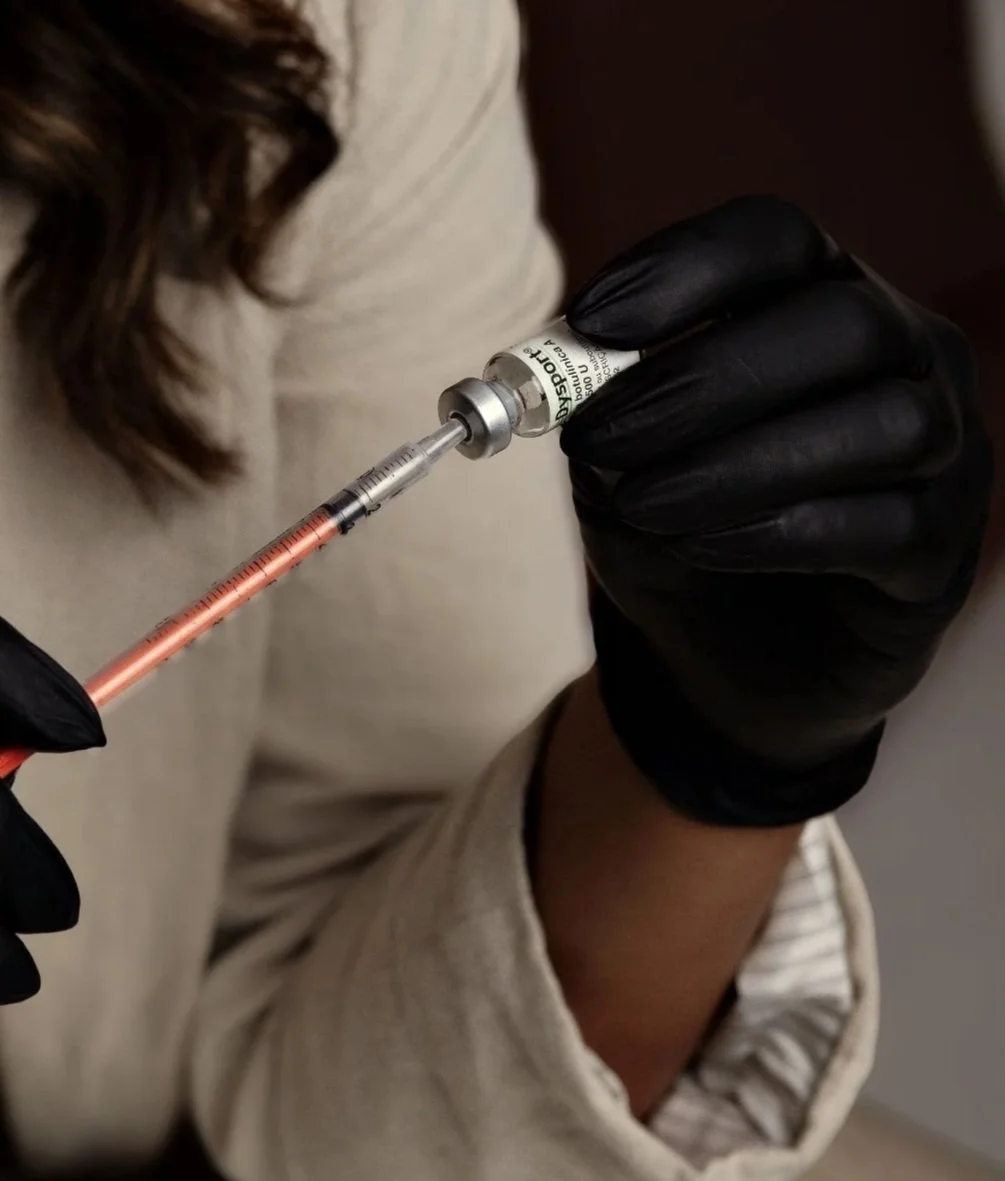 A person wearing a beige sweater and black gloves holding a syringe with a pink liquid, drawing medication from a vials.