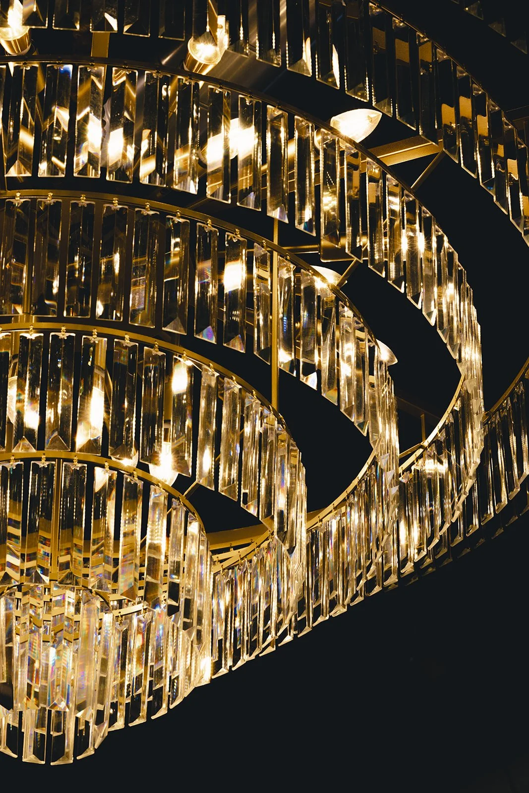 Close-up of a large, elaborate chandelier with multiple tiers of hanging, reflective gold and glass elements, glowing with warm lights.