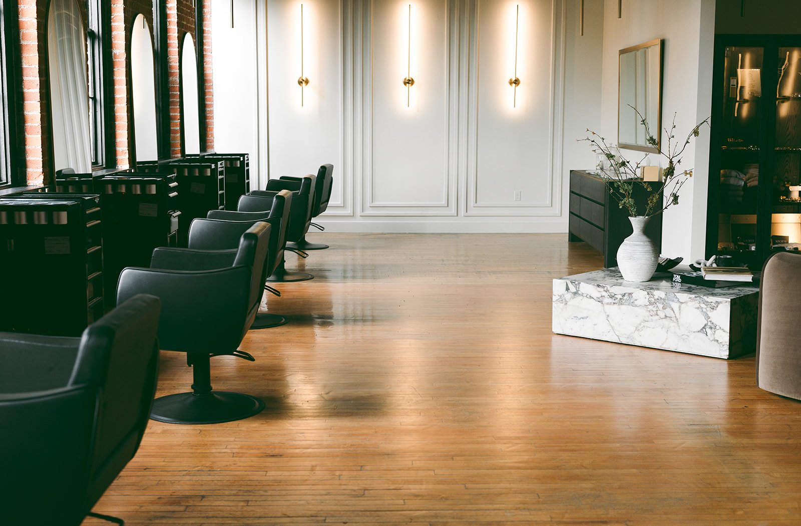 Interior of a modern hair salon or spa with black styling chairs, large windows, white paneled walls, wooden flooring, and a marble-topped side table with a vase of branches.