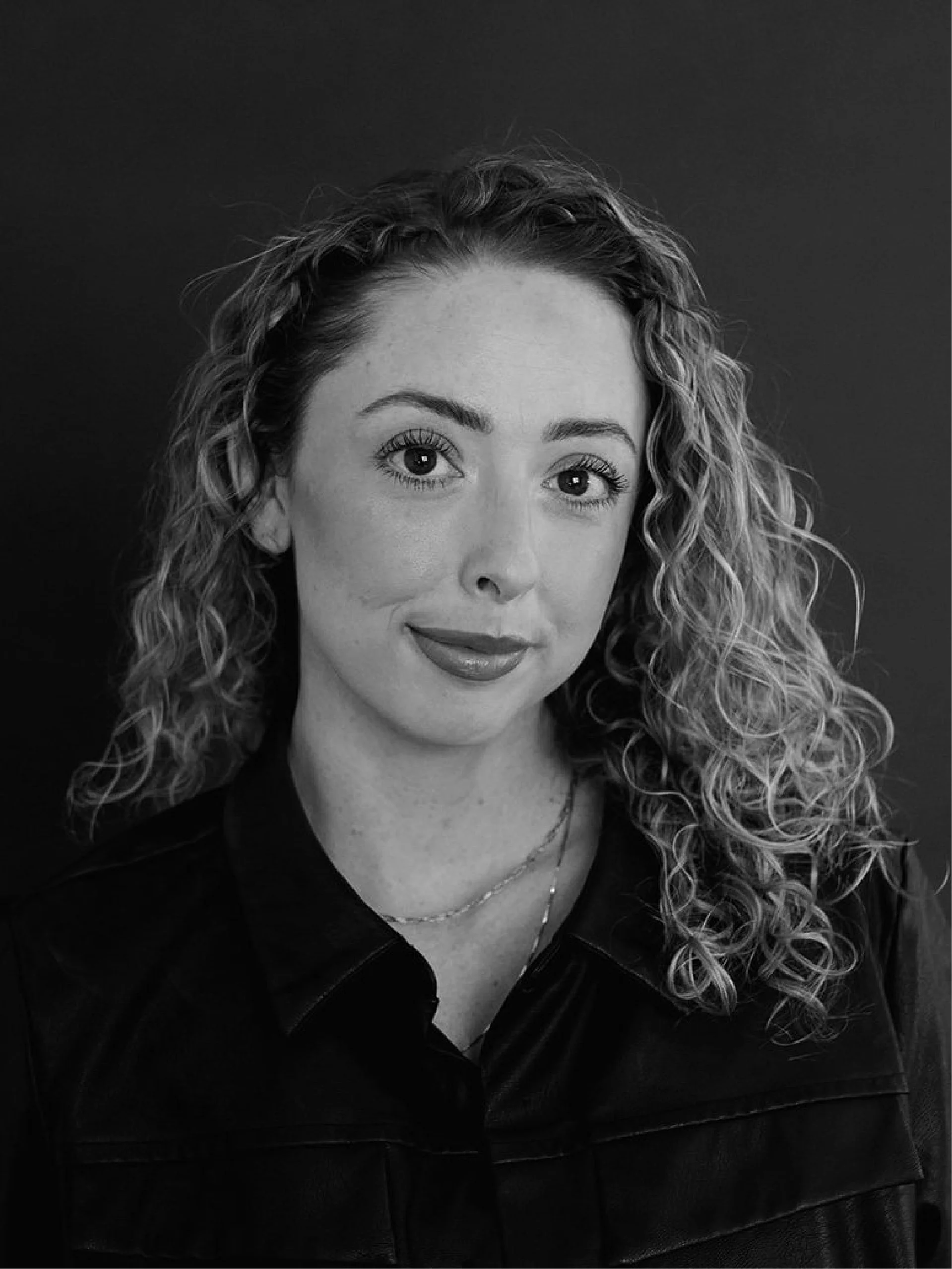 Close-up black and white portrait of a woman with curly hair, wearing a black leather jacket, a necklace, and a slight smile.