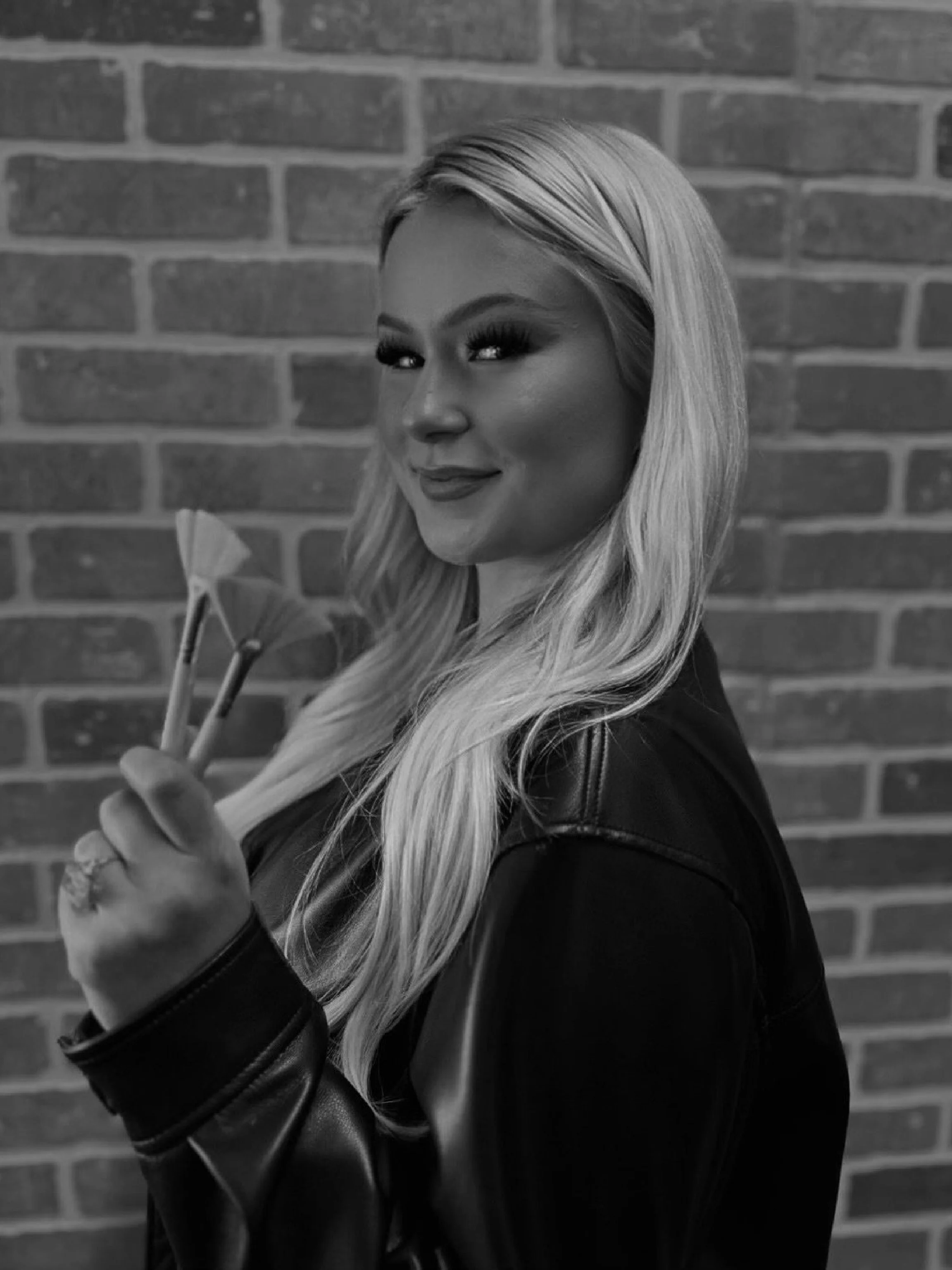 A woman with long blonde hair styled straight, wearing dark eye makeup and lipstick, holding sunglasses in one hand, standing against a brick wall.