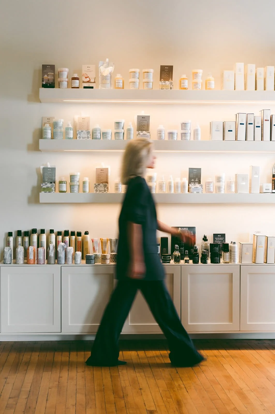 A woman with shoulder-length hair walking past shelves of skincare products in a store with wooden floors.