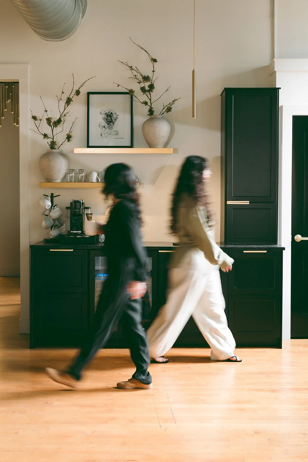 Two women walking past a black kitchen cabinet with gold handles in a modern kitchen, with decorative vases, framed artwork, and shelves in the background.