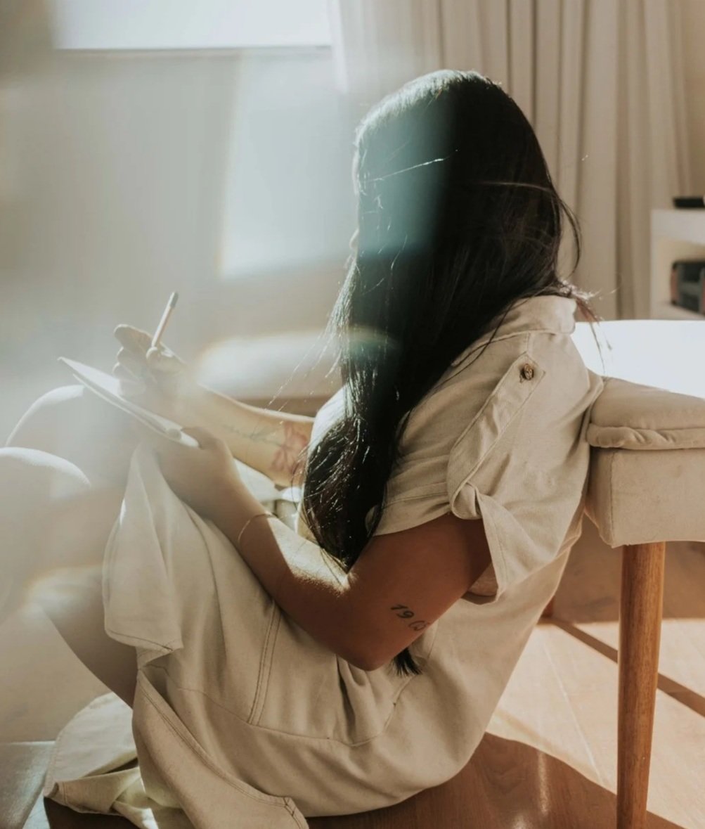 A woman with long dark hair sitting on a chair, writing in a notebook with sunlight streaming through a window.