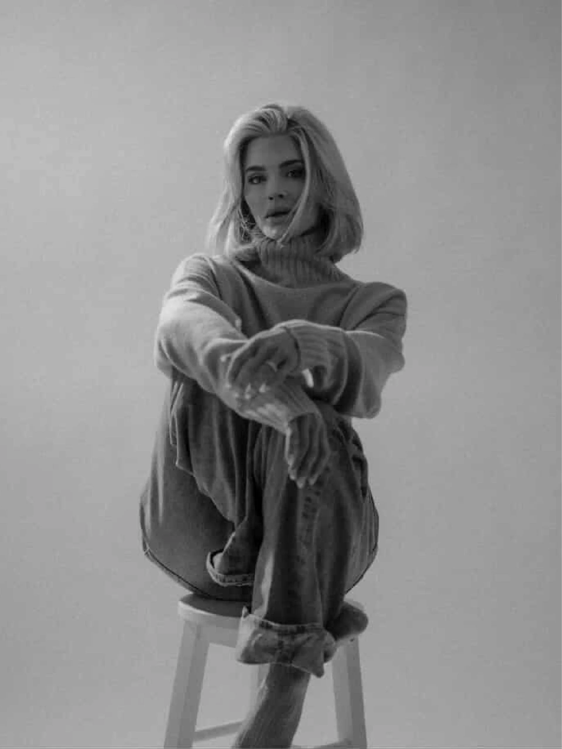 Black and white photo of a woman with shoulder-length hair sitting on a chair with crossed arms, wearing a sweater and pants, looking at the camera against a plain background.