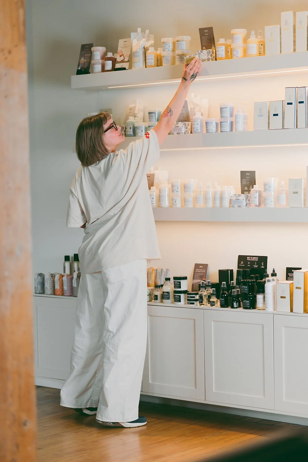 A woman with short brown hair and glasses shopping for skincare products in a store with white shelves filled with skincare bottles and boxes.