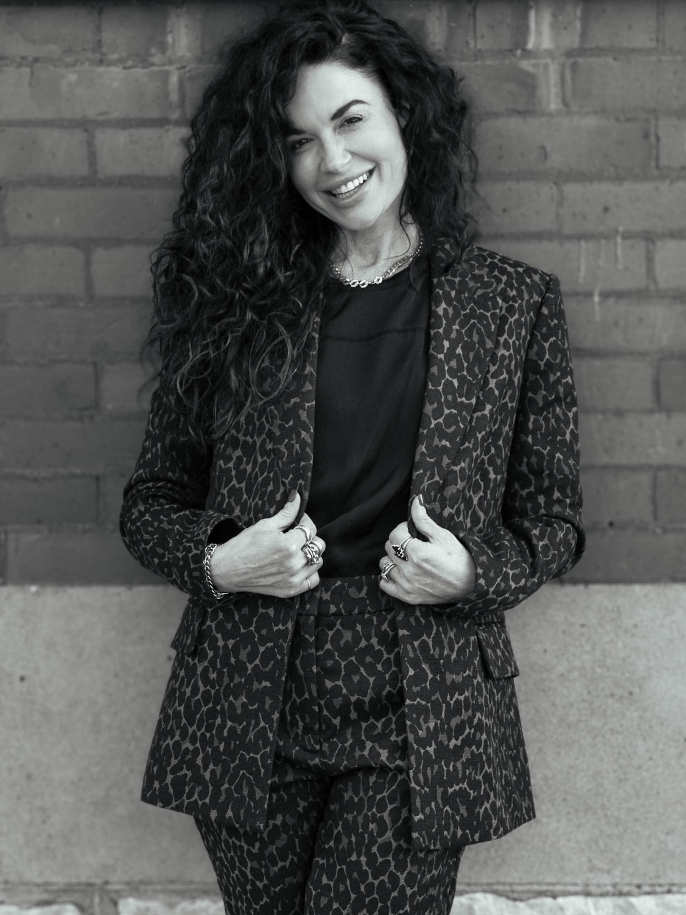 A woman with long, curly hair wearing a leopard print blazer and matching skirt, smiling and pointing at her blazer, standing against a brick wall.