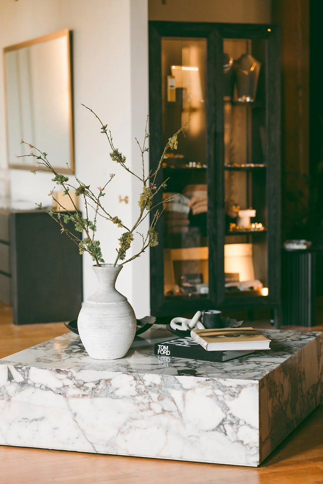 A marble table with a white vase containing dried branches, a black book, and a white chain link sculpture inside a modern interior.