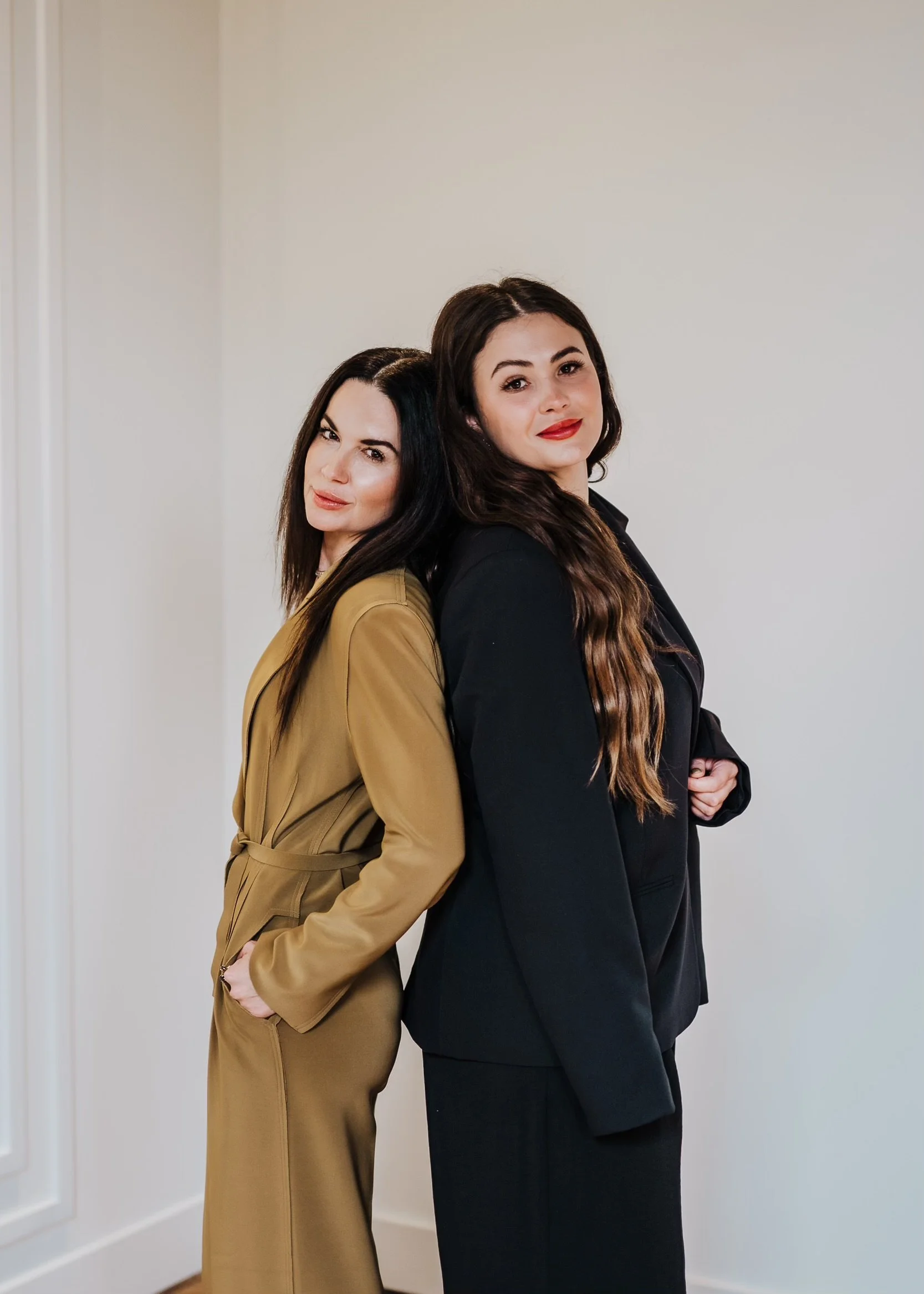 Two women are standing back to back, leaning against each other, in front of a plain beige wall. One woman is wearing a tan trench coat, and the other is wearing a black blazer.