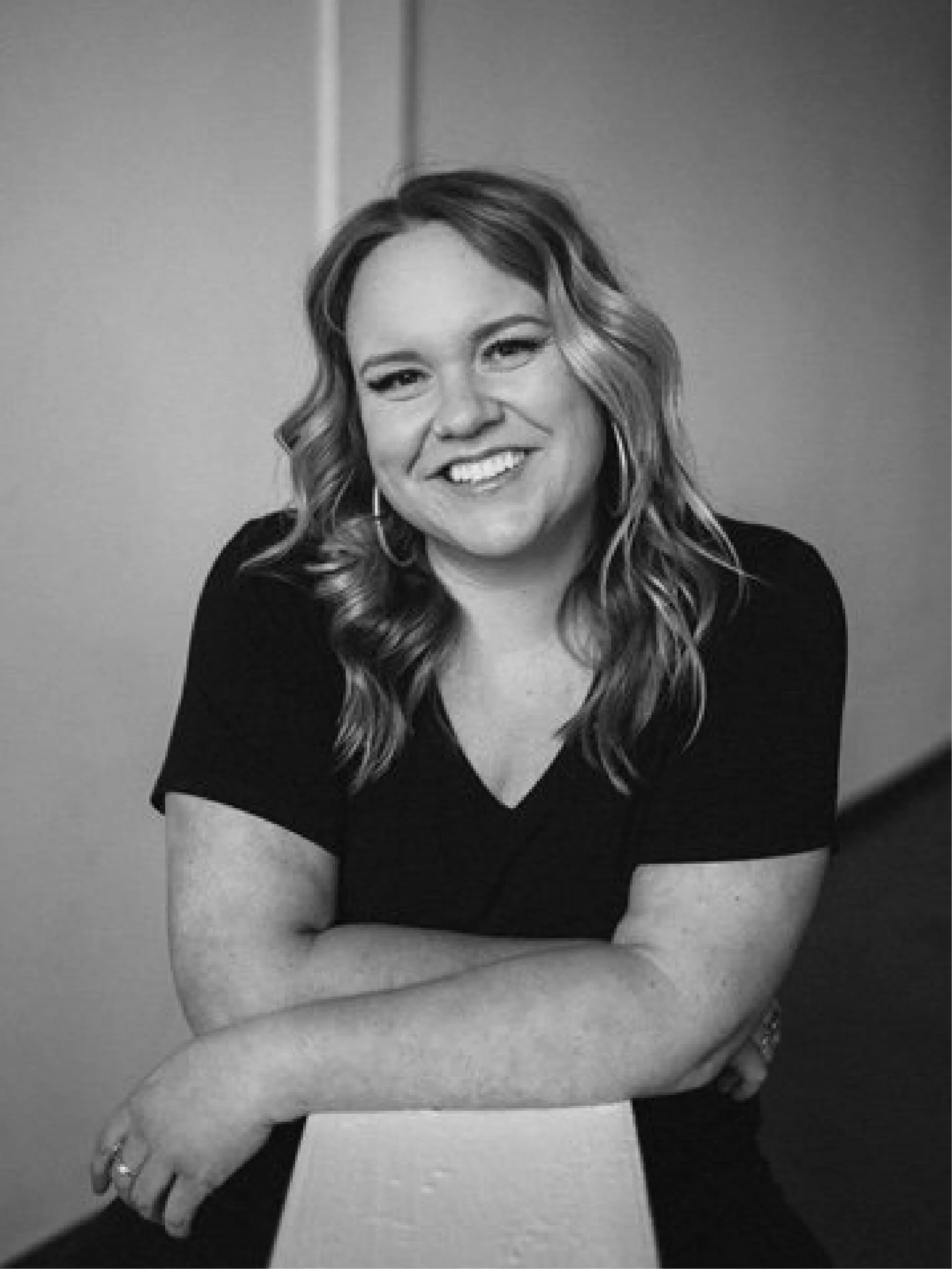 Black and white photo of a woman smiling with arms crossed, wearing earrings and a short sleeve top.