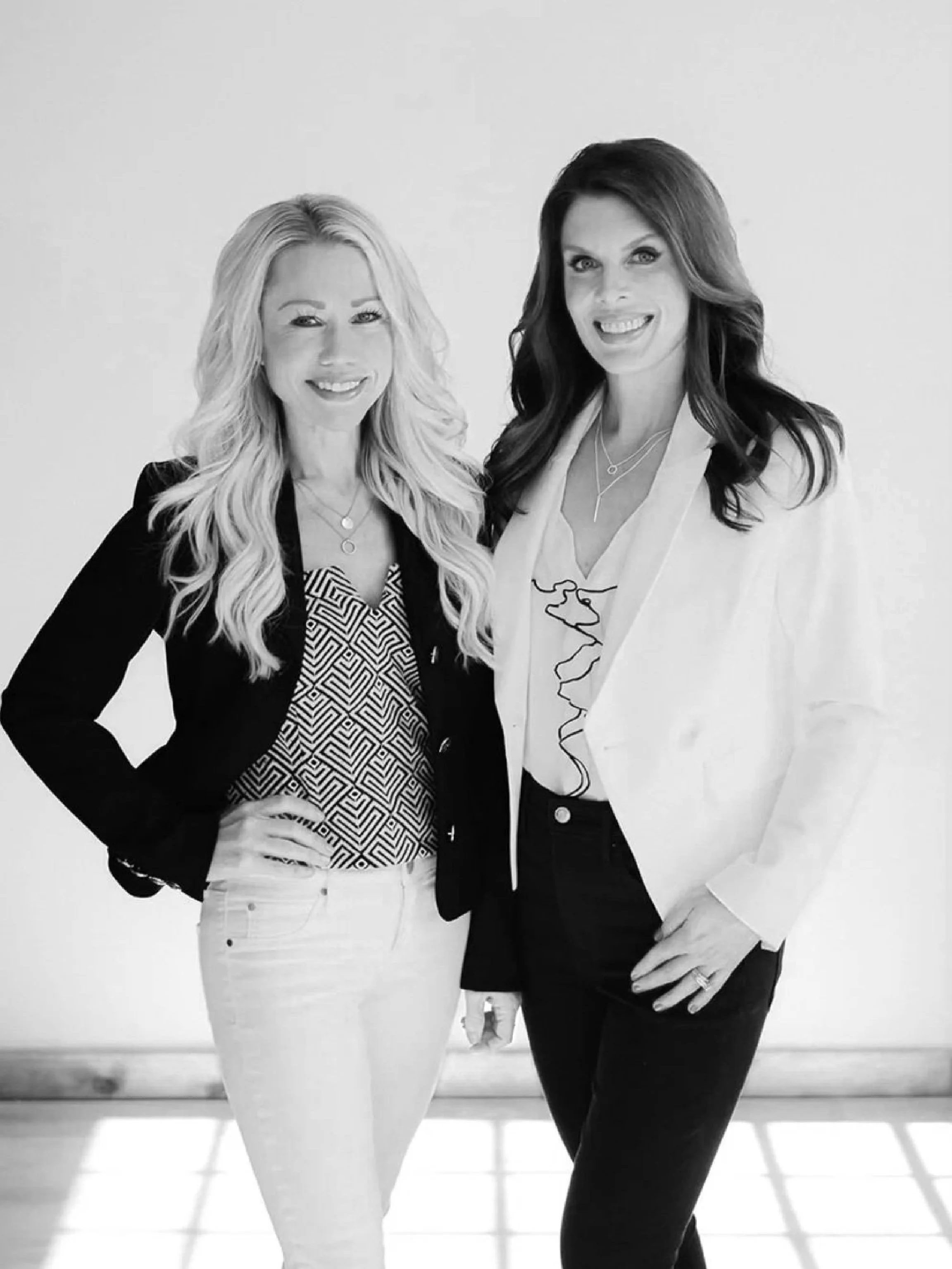 Two women standing together smiling with one arm around each other's waist, in front of a plain wall, in a black-and-white photograph.